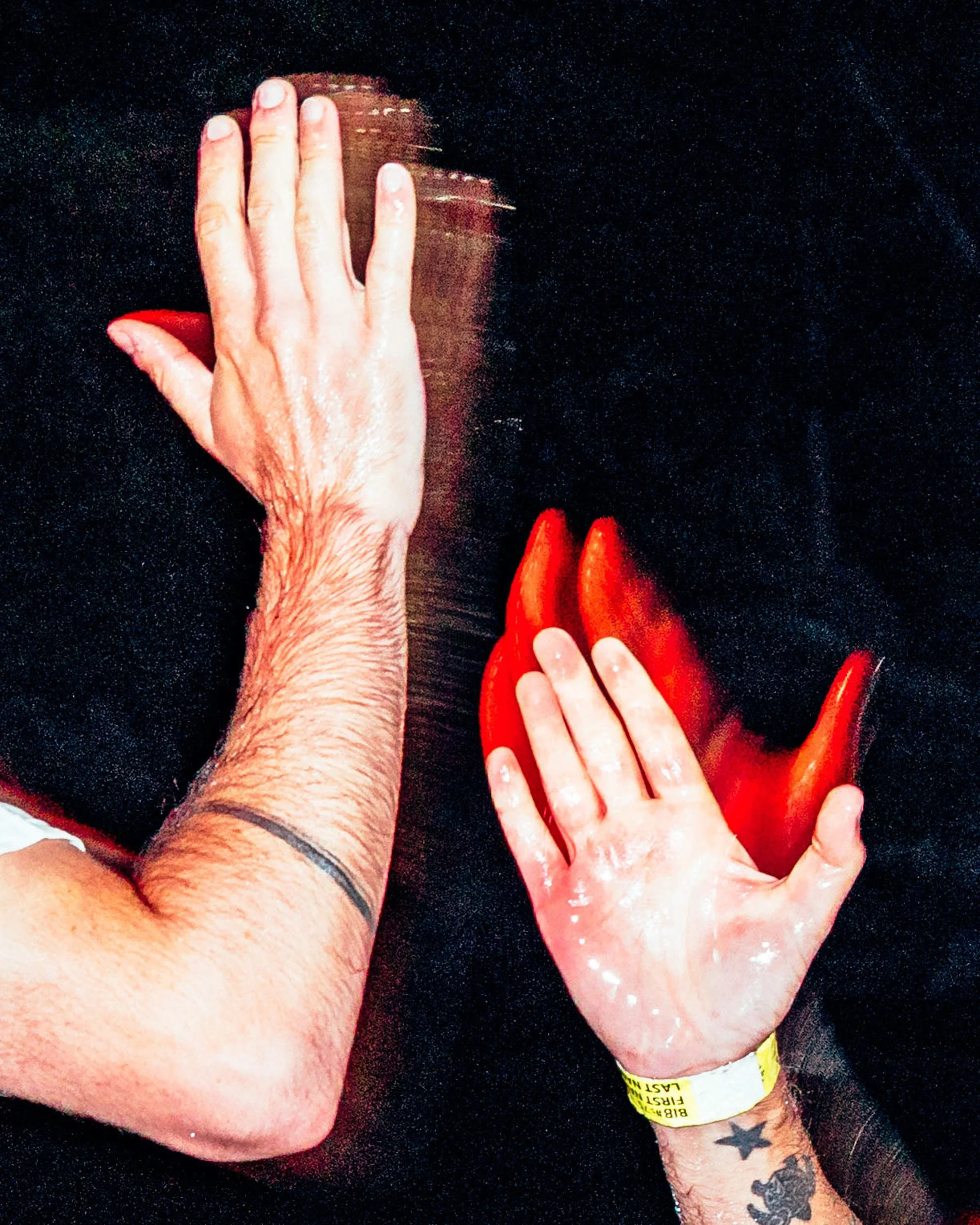 Multiple hands and forearms with tattoos touching a black surface. One hand is holding a red chili pepper, and one hand is covering the face of a person wearing a dark shirt. The image has a dark background with small light-colored specks.