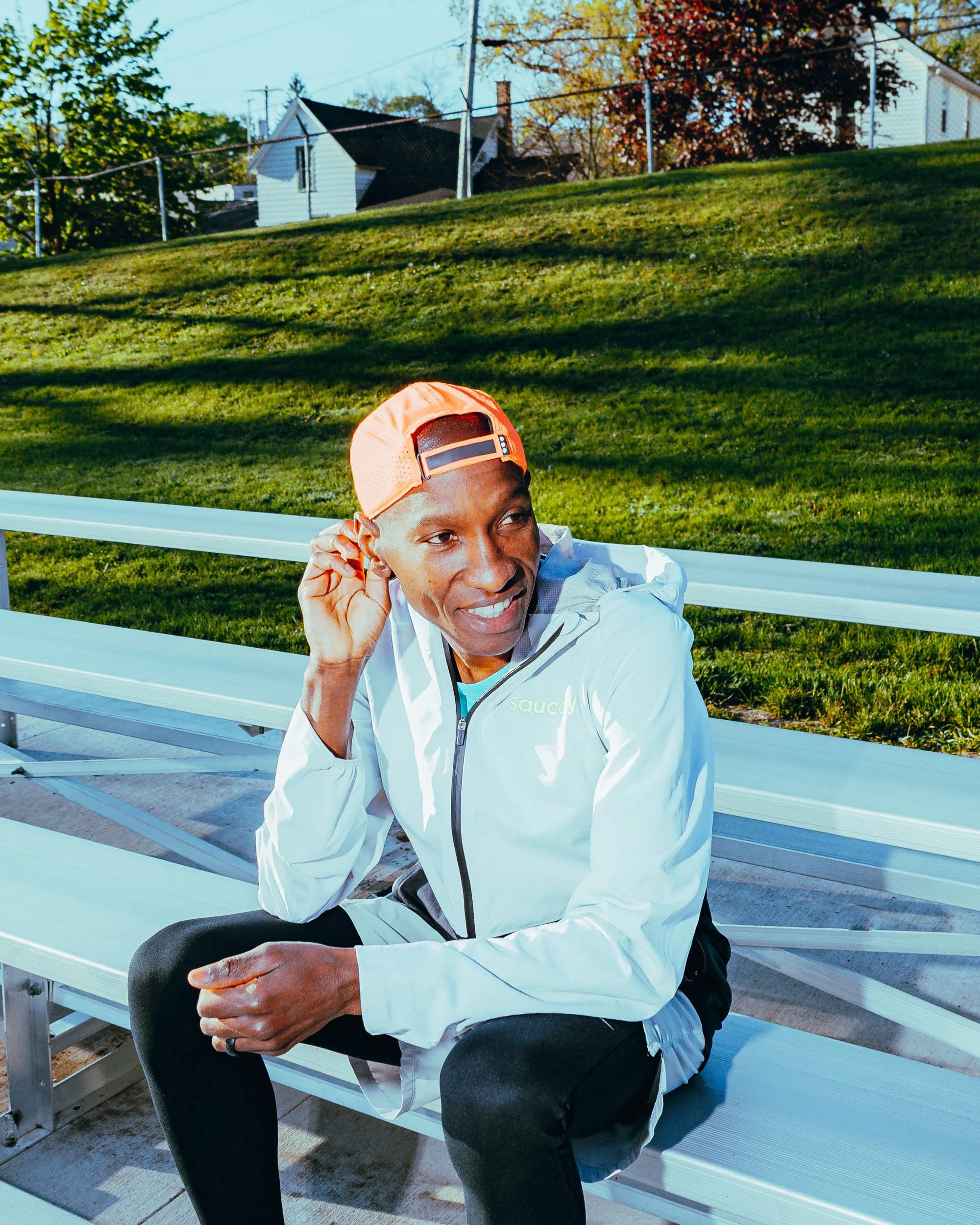 A man sitting on a white bench outdoors, wearing a white jacket, black pants, and an orange cap, smiling and holding his glasses with his right hand. The background features a grassy hill and residential houses with trees.