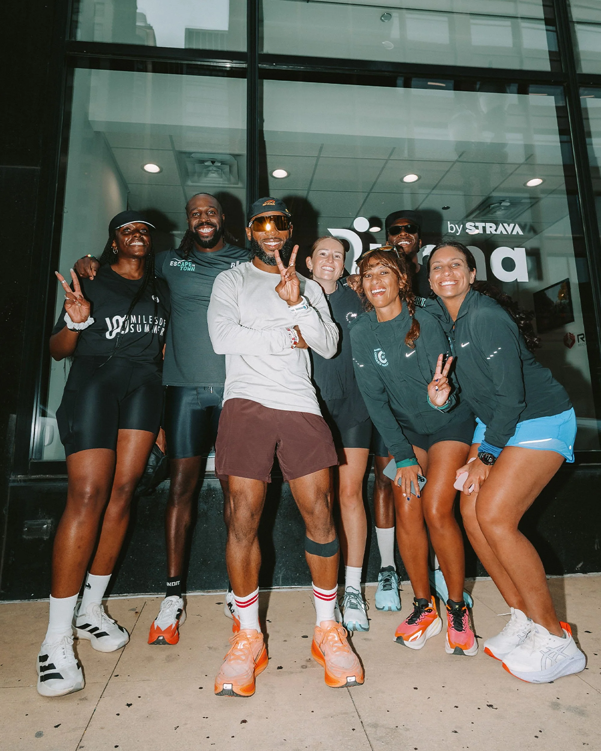 A group of seven diverse people standing together outside a building, smiling and posing for a photo, wearing athletic gear and sneakers, with some raising peace signs.