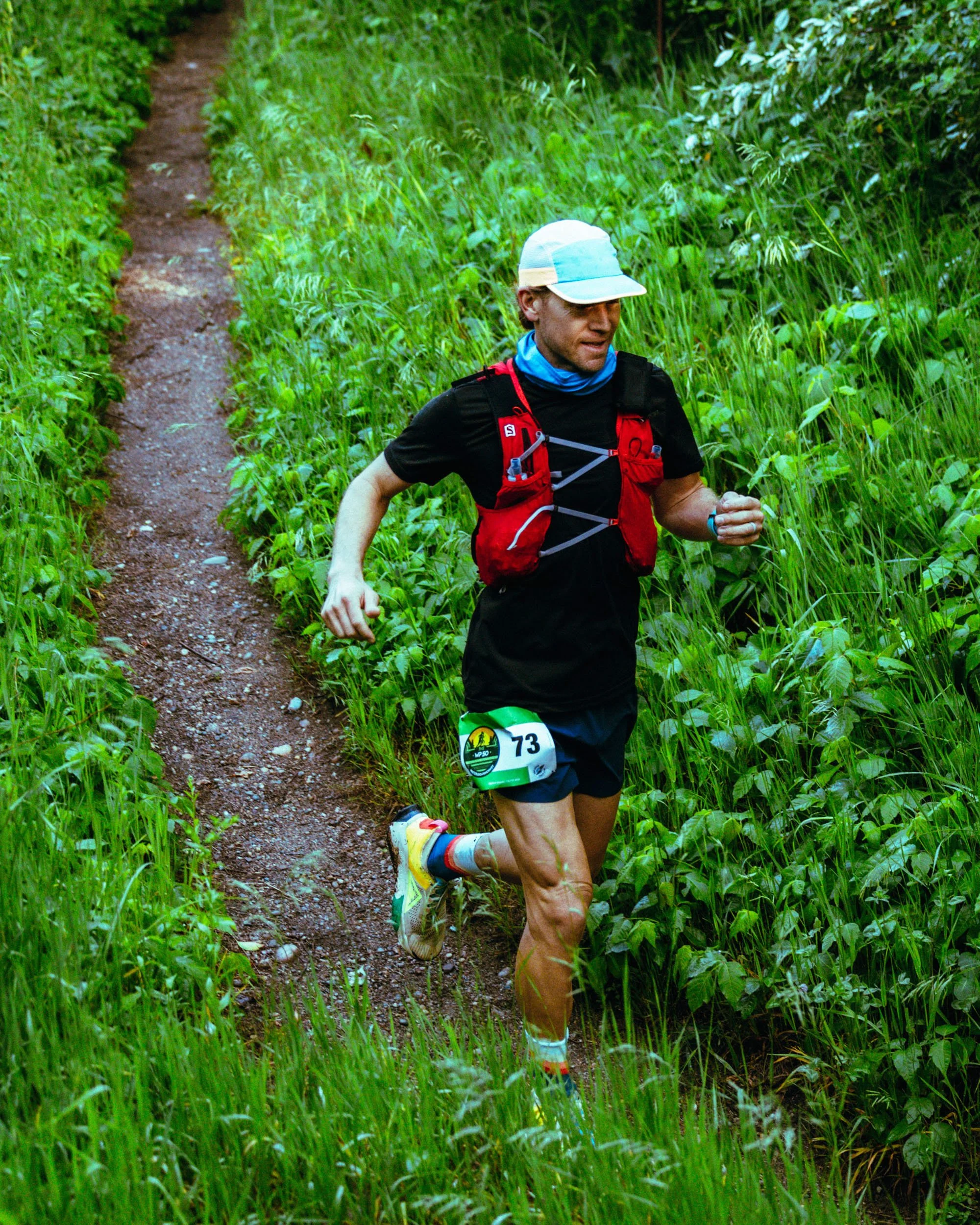 Male runner wearing a black T-shirt, shorts, and a colorful hat, running on a narrow dirt trail surrounded by green grass and tall plants in a lush outdoor setting.