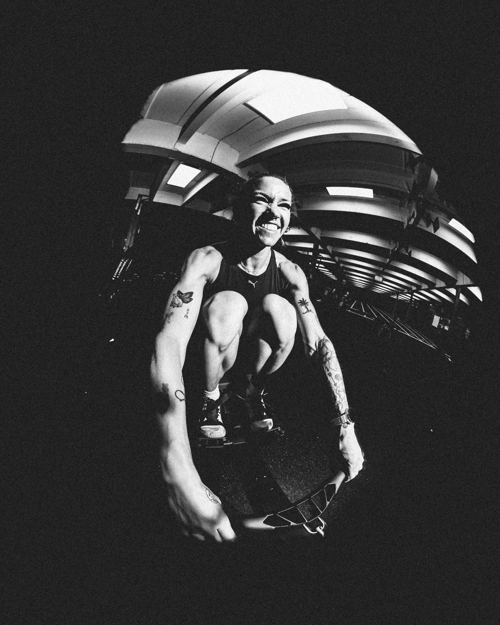 A black-and-white photo of a woman with tattoos crouching next to a small trampoline, smiling widely, with a modern architectural structure in the background.