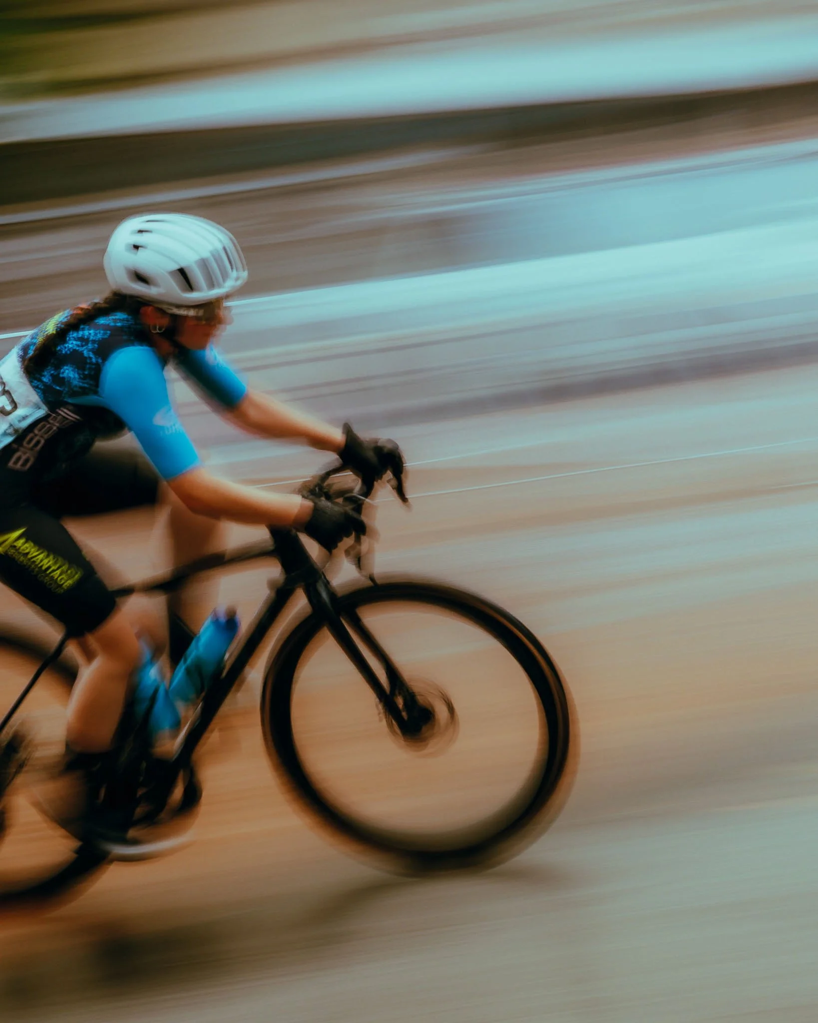 A cyclist wearing a white helmet and blue cycling gear riding a black bicycle on a wooden indoor track in motion, with a blurred background.