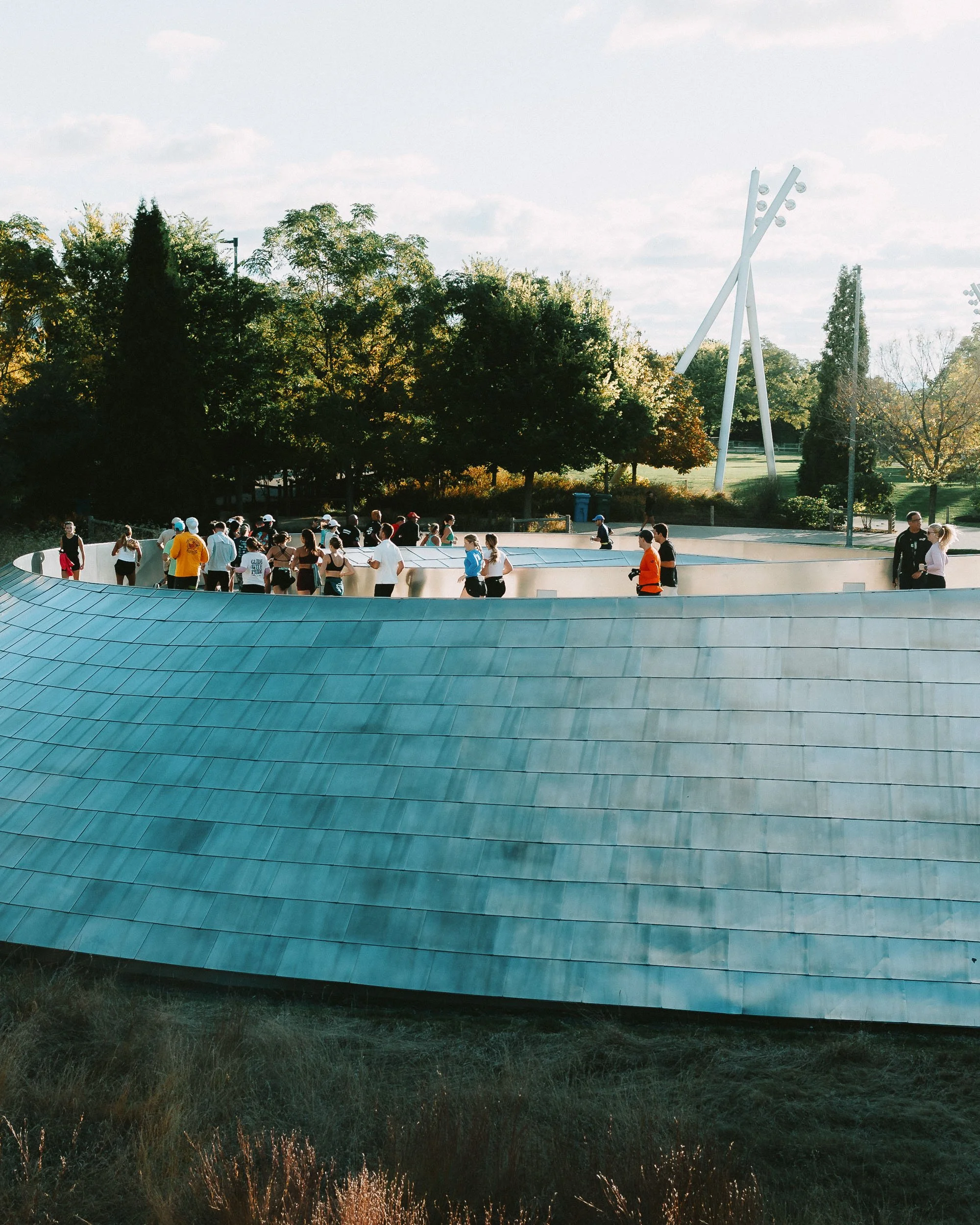 Large modern architectural building with a curved, teal-colored roof, adjacent to a green park with trees and tall light poles, under a partly cloudy sky; people walking and gathering outside.