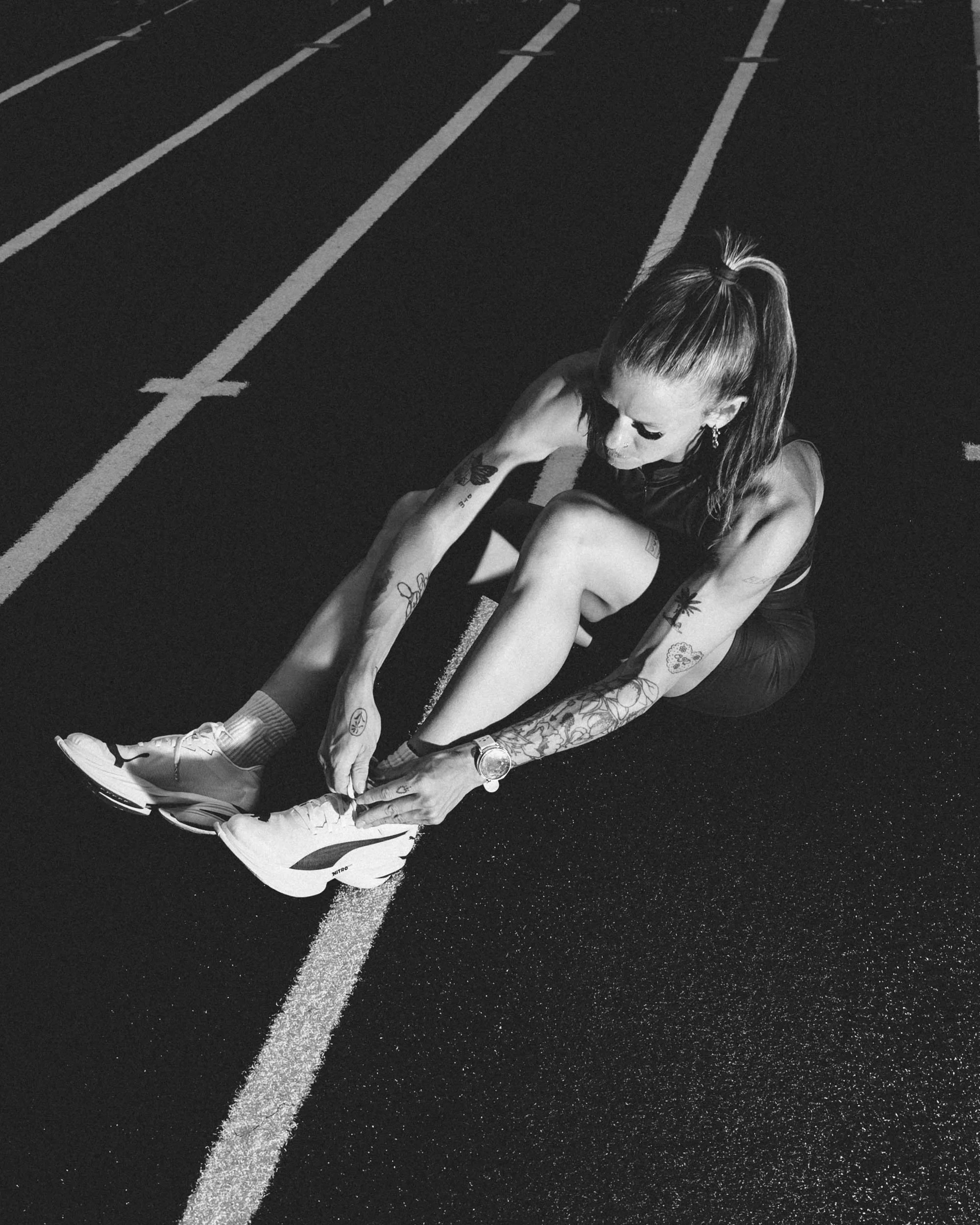 A woman with tattoos on her arms and long hair tied in a ponytail is sitting on a running track, tying her shoelaces. She wears athletic clothing and sneakers, with sneakers on her feet. The photo is in black and white.