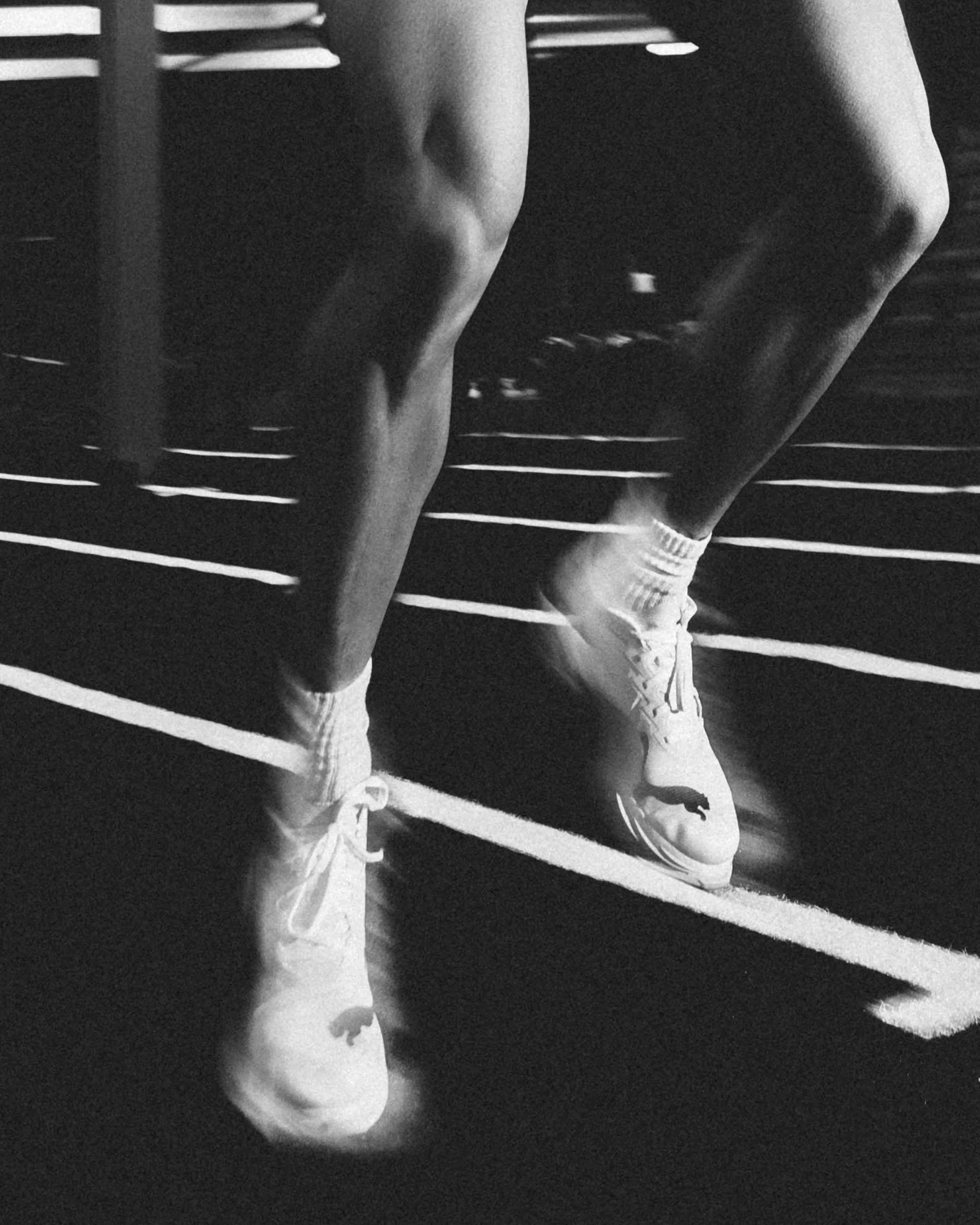 A black and white photo of a person's legs, wearing sneakers and socks, standing on a striped running track.