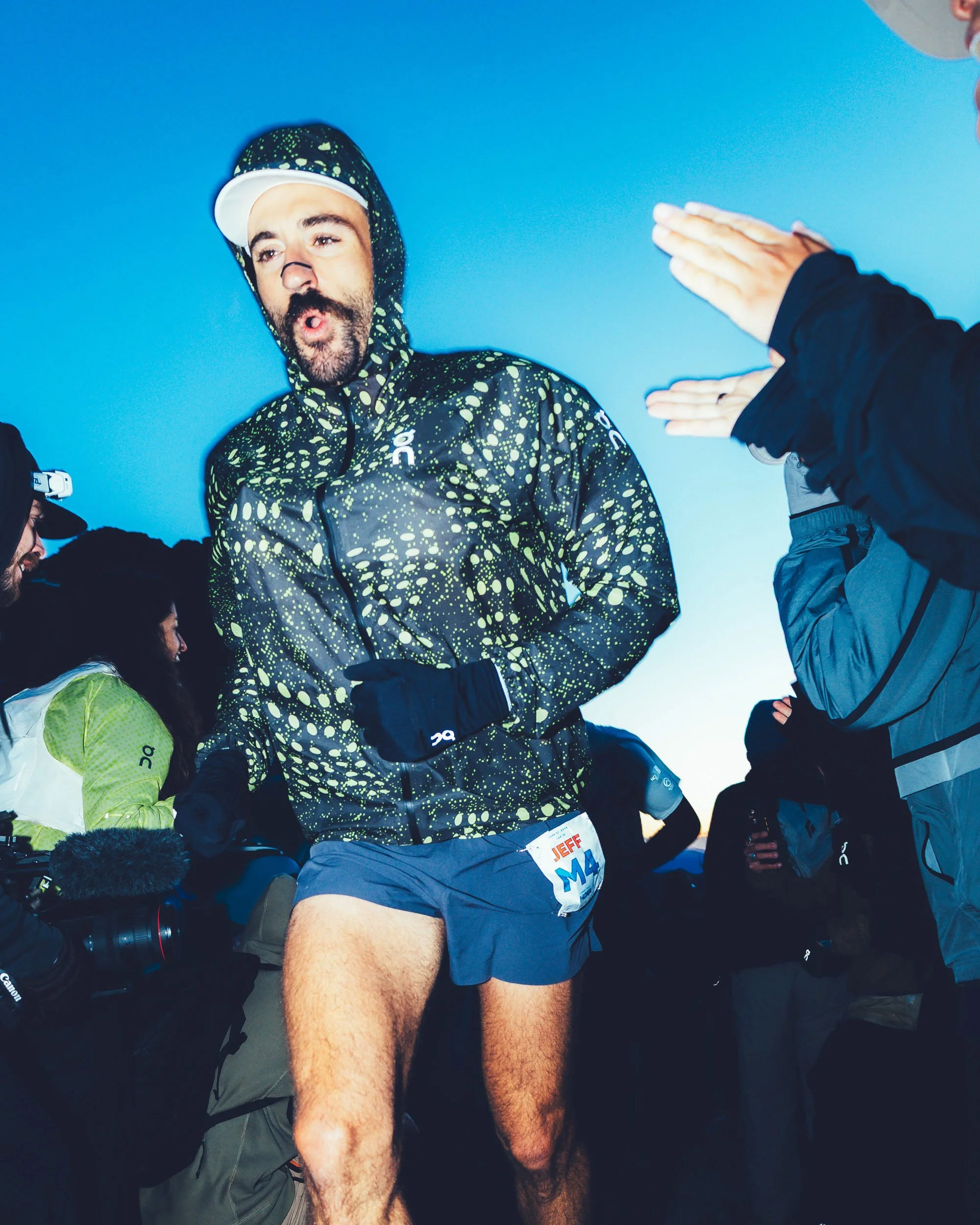 A man with a beard and mustache wearing a black and yellow patterned jacket, a white cap, and running shorts, appears to be in a race or similar event, surrounded by other people.