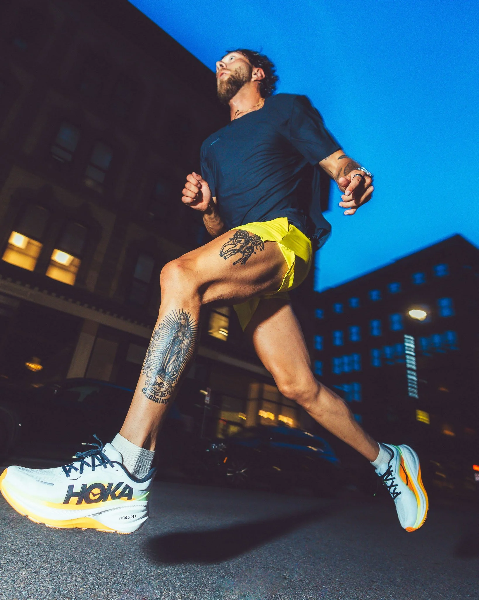A man running outdoors at night in an urban setting, wearing white athletic shoes, yellow shorts, a black t-shirt, and equipped with a watch and tattoos on his arms and leg.
