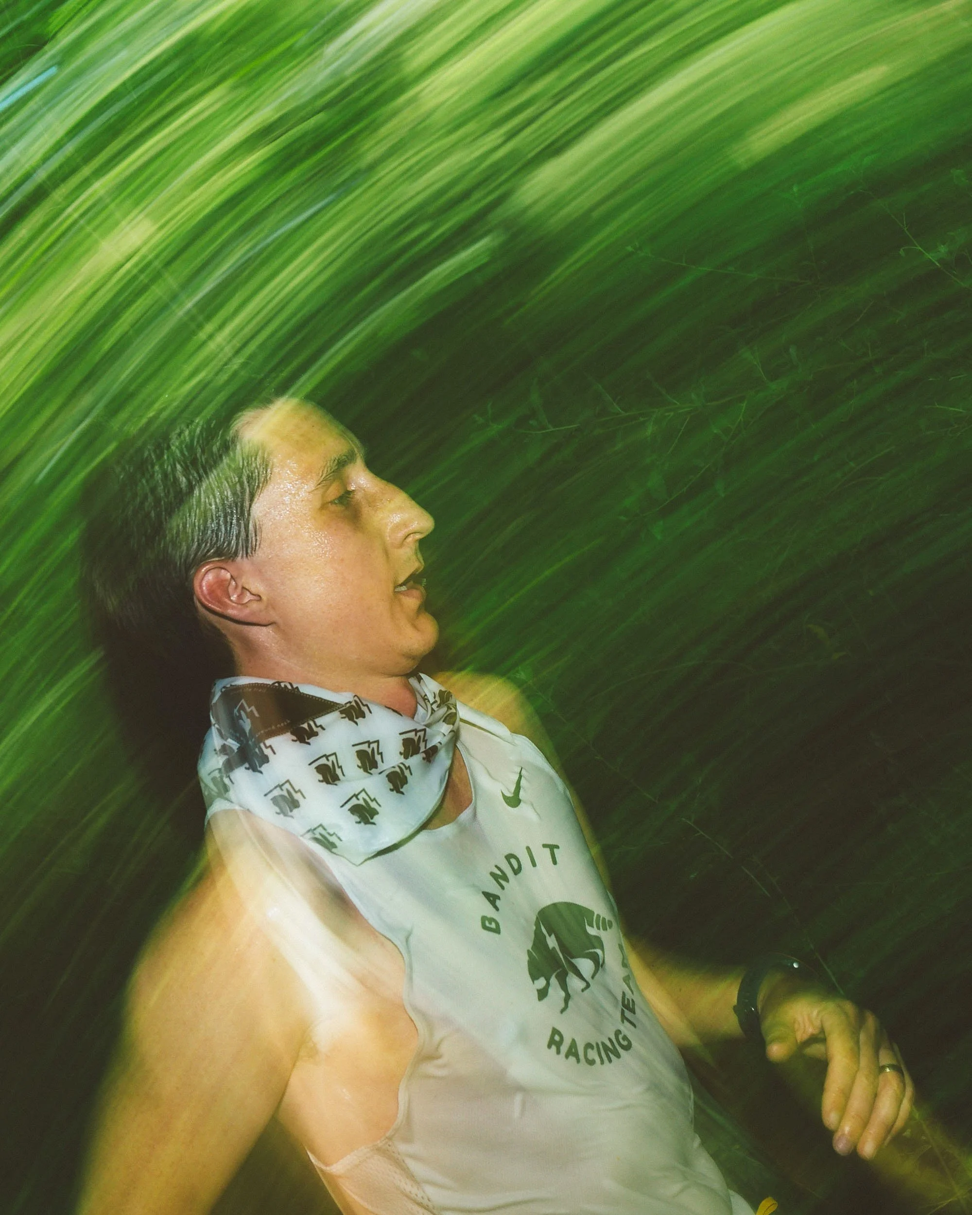 A person with wet hair, wearing a white shirt and a white bandana with black lettering, sitting in a natural setting surrounded by tall green grasses.