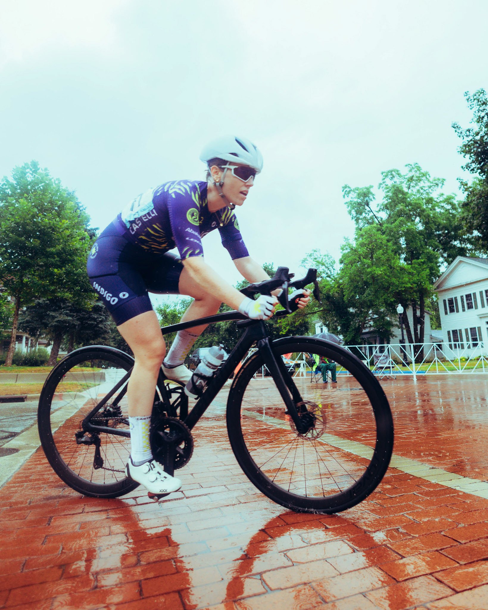 A cyclist wearing a purple and black cycling outfit, a white helmet, and sunglasses rides a black bike on a wet brick pathway during rainy weather. Green trees and white buildings are visible in the background.