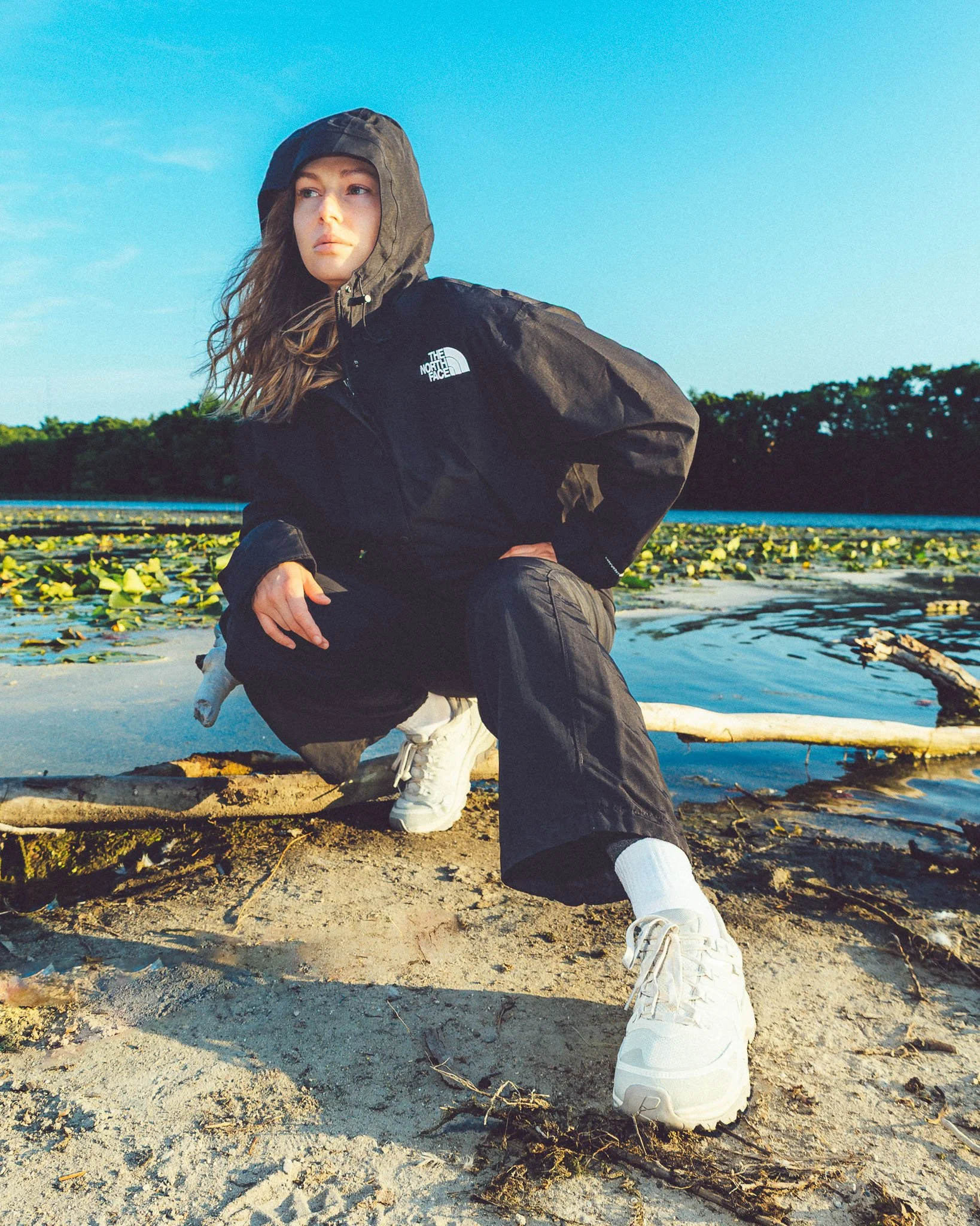 A young woman wearing a black North Face jacket, kneeling on a sandy shore with water and green trees in the background. She is looking to her left with her hood up and her right hand on her knee, with her left hand resting on her thigh. She is weari