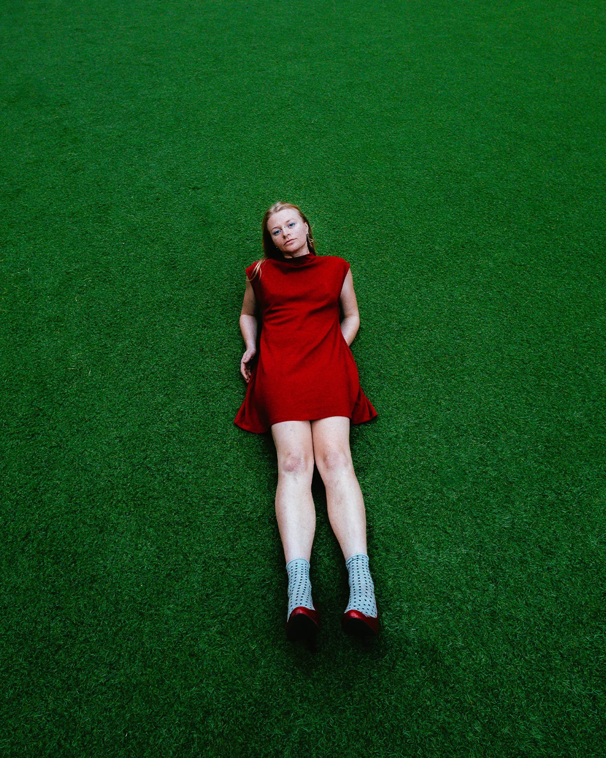 A young woman with red hair laying on a green grass field, wearing a red dress, white socks with black dots, and red shoes.
