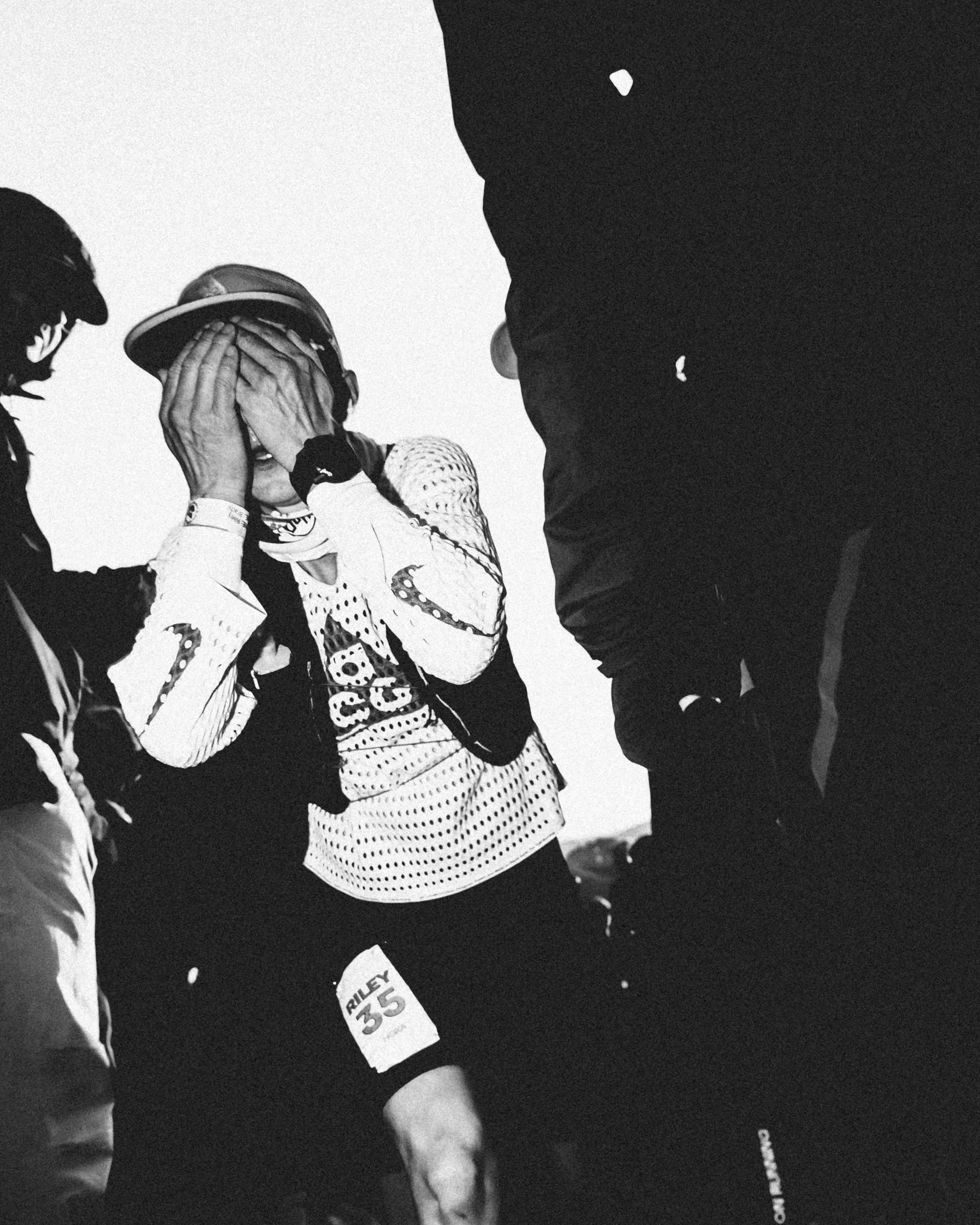 A person dressed in racing gear with a racing number 35, covering their face with both hands, standing among other people at what appears to be a racing event.