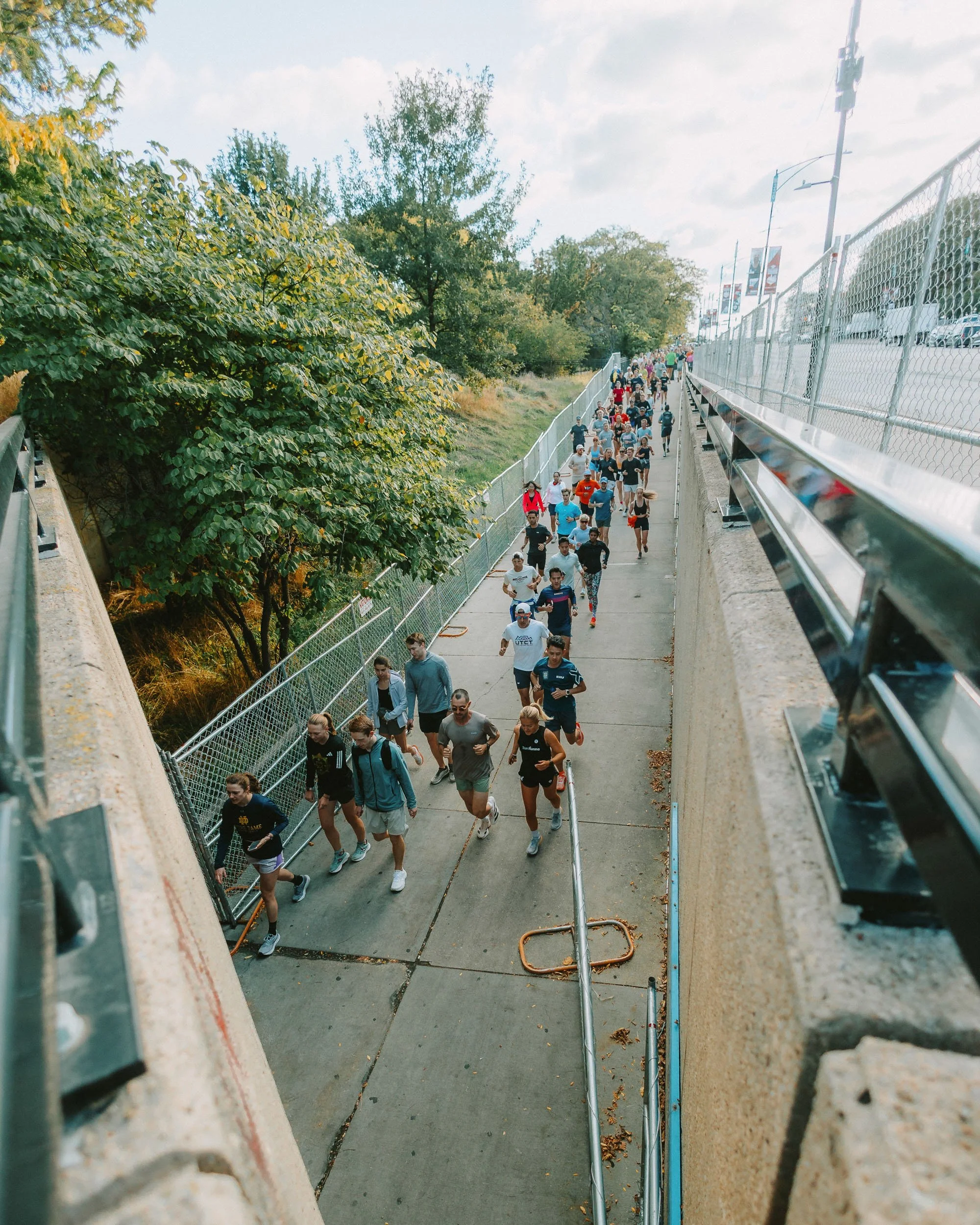 A large group of people running or walking on a concrete sidewalk next to a chain-link fence, with trees and a cloudy sky in the background. The photo is taken from an elevated perspective.