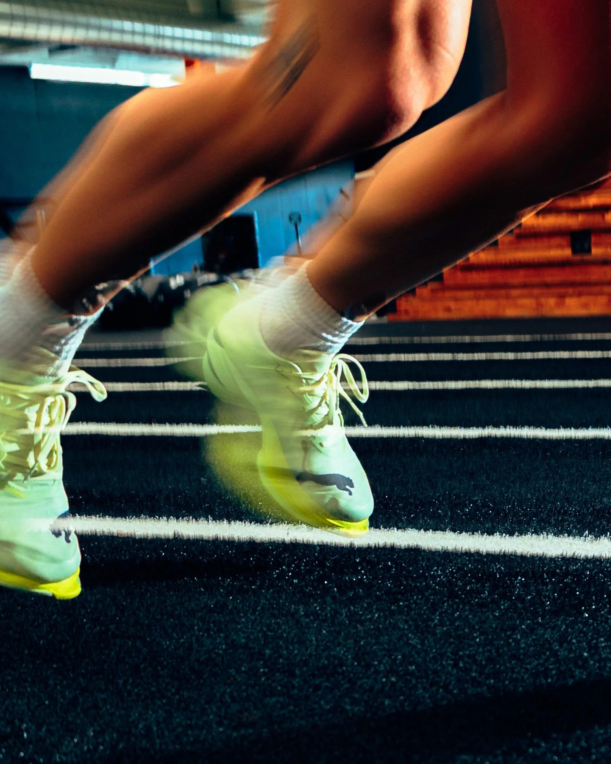 Close-up of a person wearing yellow running shoes, white socks, and workout shorts, crouching on a black textured track surface, with wooden bleachers and a blue building in the background.