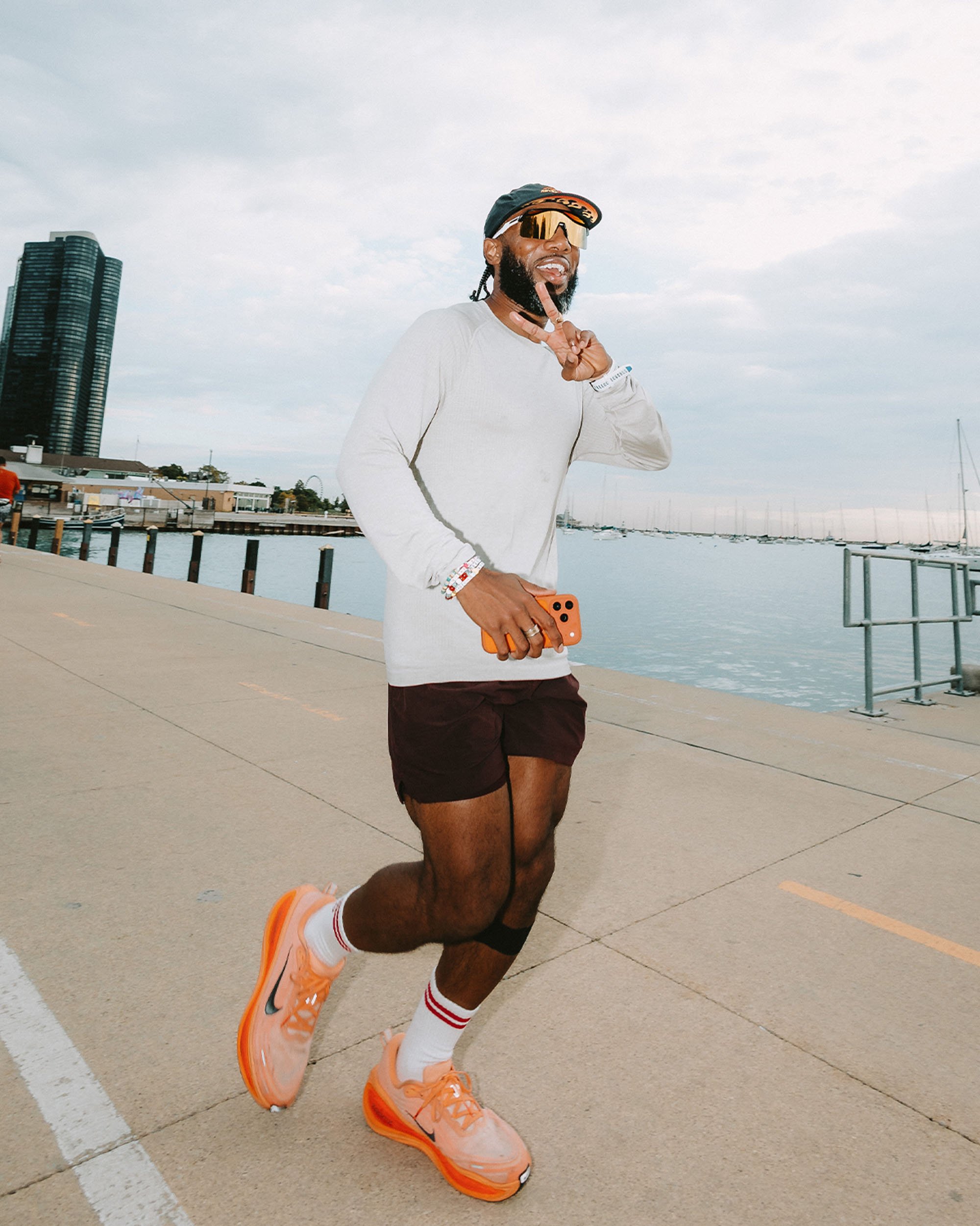 A man jogging outdoors near a waterfront with sailboats, wearing orange sneakers, white socks with red stripes, and a white shirt, holding a phone, and making a peace sign with his fingers.
