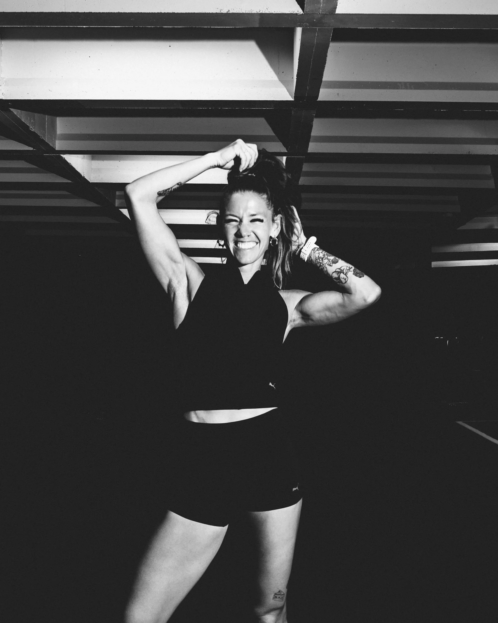 Black and white photo of a woman with tattoos, smiling, wearing a sleeveless top, pulling her hair up with one hand, underneath a structure with horizontal beams.