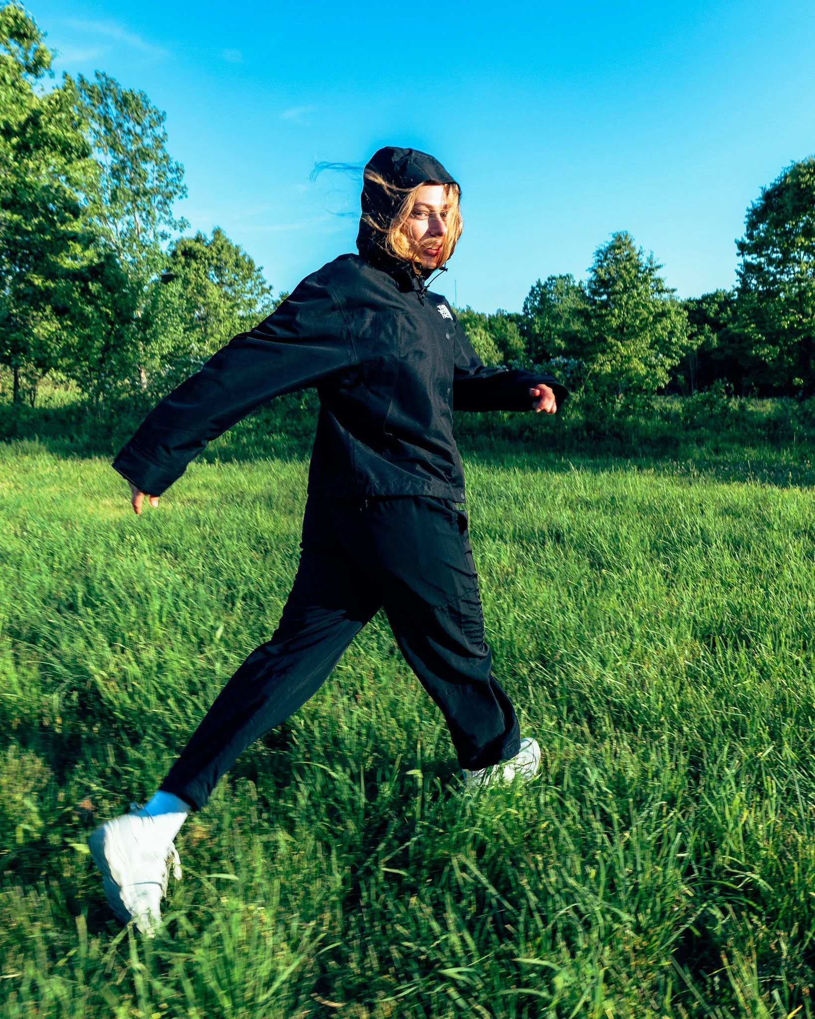 A person wearing a black North Face jacket with a hood and black pants walking through a green grassy field with trees in the background under a clear blue sky.