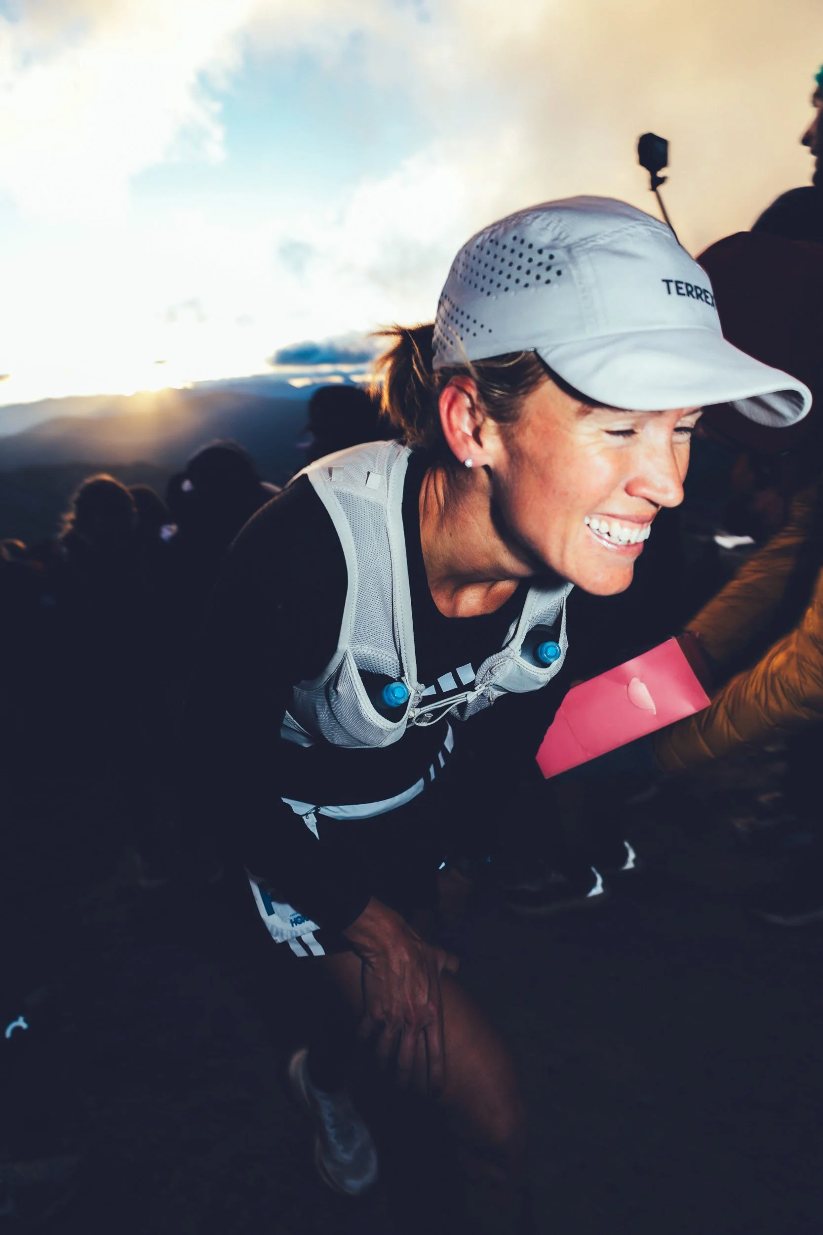 Close-up of a woman smiling during a race, wearing a white cap, black athletic clothing, and a hydration vest, with a sunset or sunrise in the background.