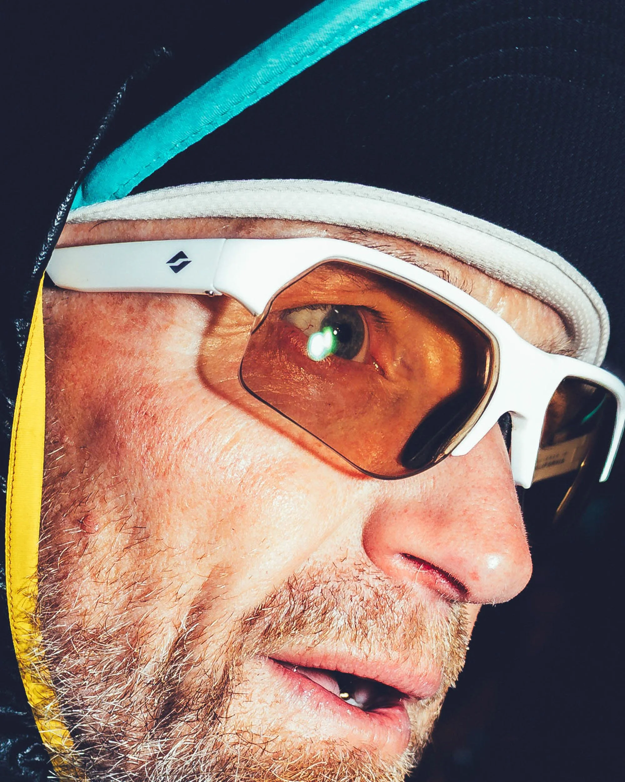 Close-up of a man's face wearing sunglasses and a winter hat, with visible facial hair, and a reflection of a green object in the sunglasses.