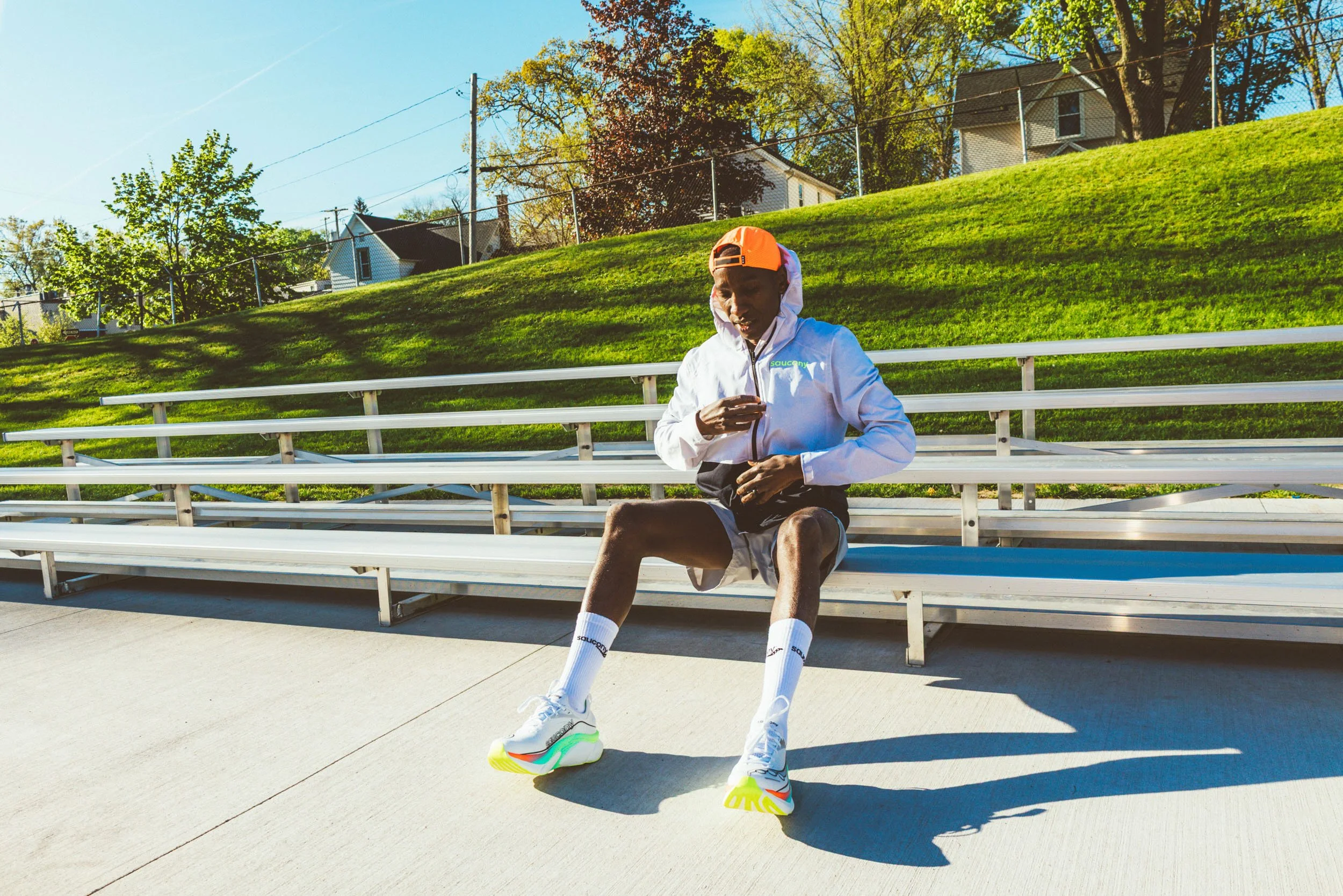 A man sitting on bleachers at a park, dressed in sportswear including a white jacket, shorts, and colorful running shoes, looking at his phone with trees and houses in the background on a sunny day.