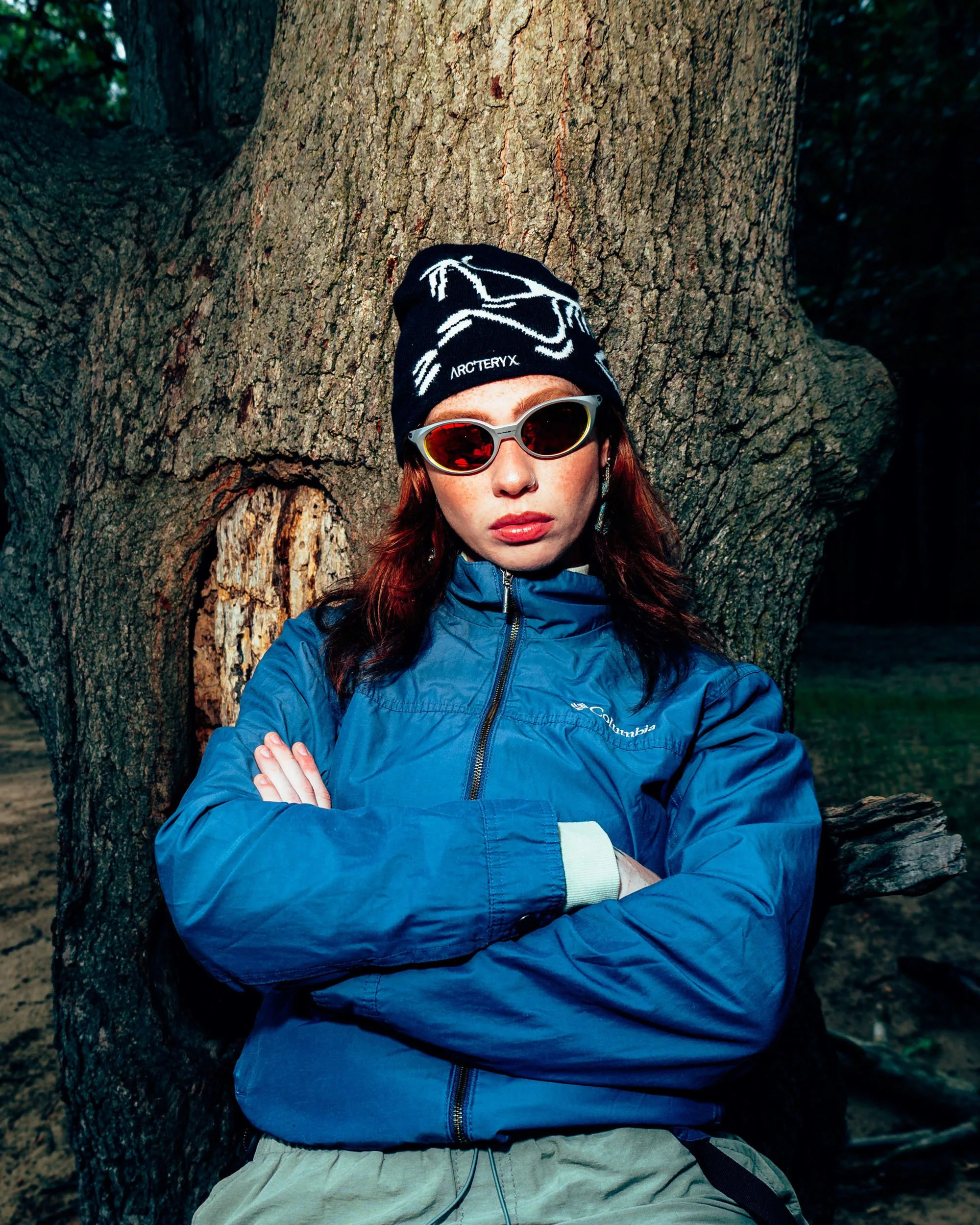 A woman with red hair wearing a black beanie, red-tinted sunglasses, a blue Columbia jacket, and beige pants poses with crossed arms in front of a large tree trunk.