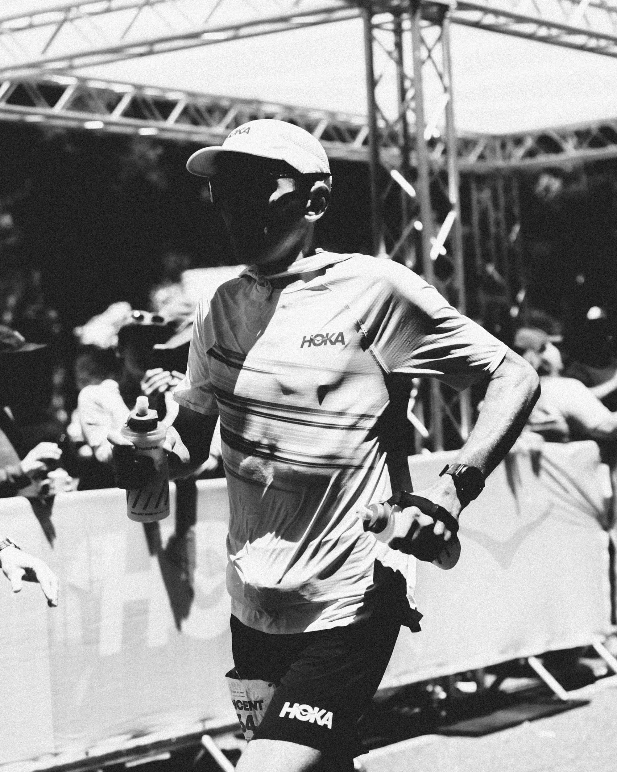 A man in athletic gear, wearing a hat and sunglasses, is running outdoors in a competitive event, with a crowd and stage equipment in the background.