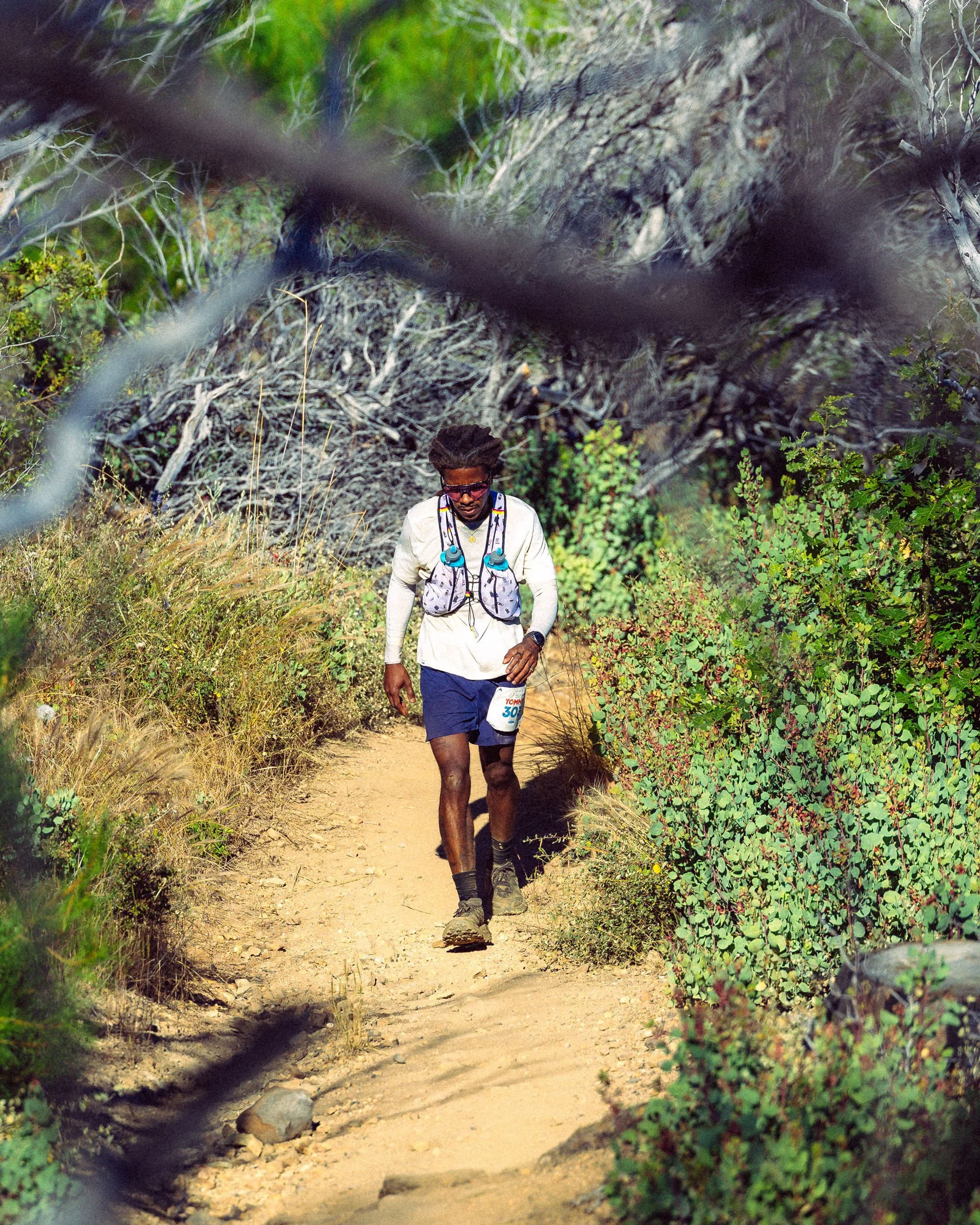 A man hiking on a dirt trail in a lush, green outdoor setting, wearing a white long-sleeve shirt, dark shorts, hiking shoes, and sunglasses, with a hydration pack and race bib number 30.