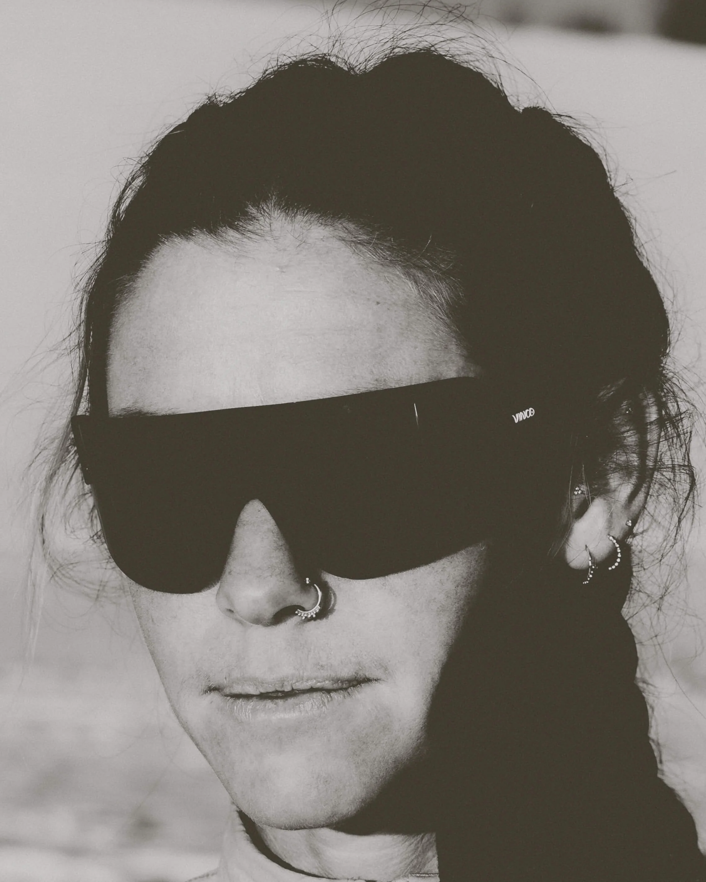 A black-and-white close-up portrait photograph of a woman with dark hair, wearing hoop earrings, and dark sunglasses. She has a septum piercing and glossy lips.