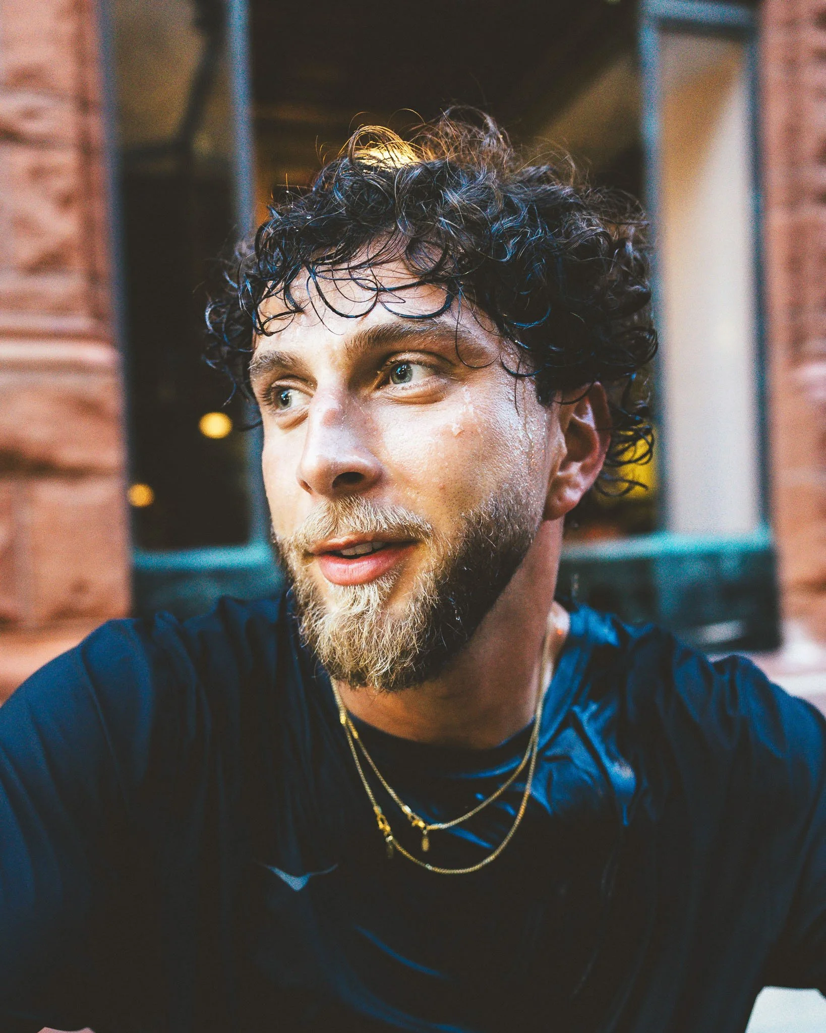 Close-up of a man with curly dark hair, light skin, wearing multiple gold necklaces and a dark shirt, with a background featuring brick wall and blurred lights.