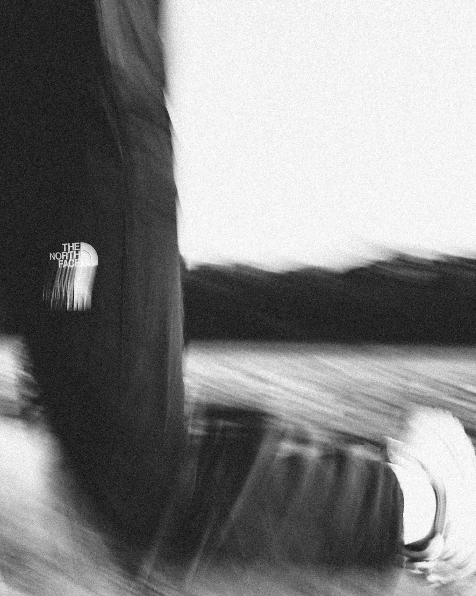 Close-up black and white photo of a person's hand holding a dark object, possibly a notebook or a book, with a white logo that says 'The North Face'. The background is blurry with a swirling pattern.