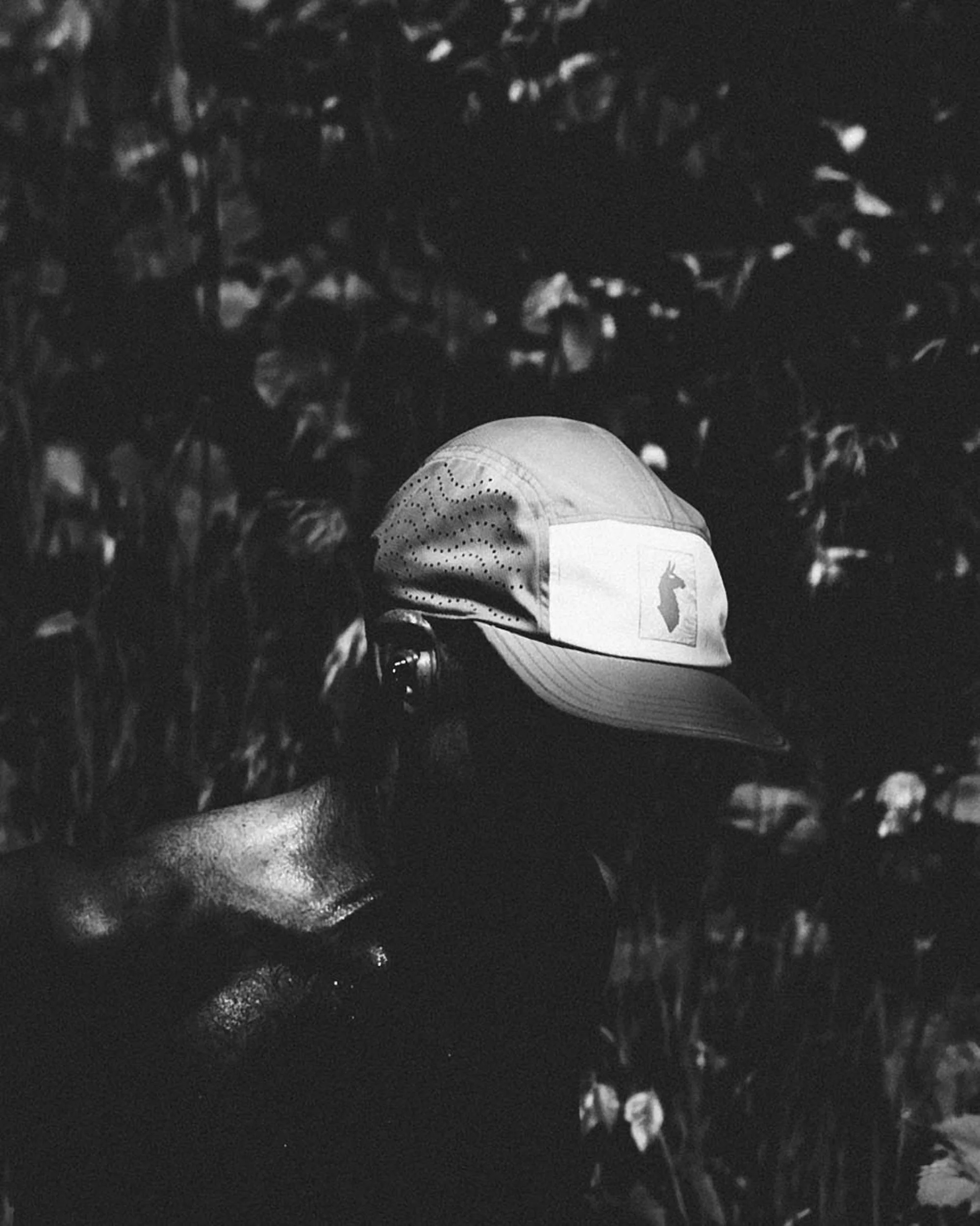 Black and white photograph of a person wearing a baseball cap, with trees and foliage in the background.