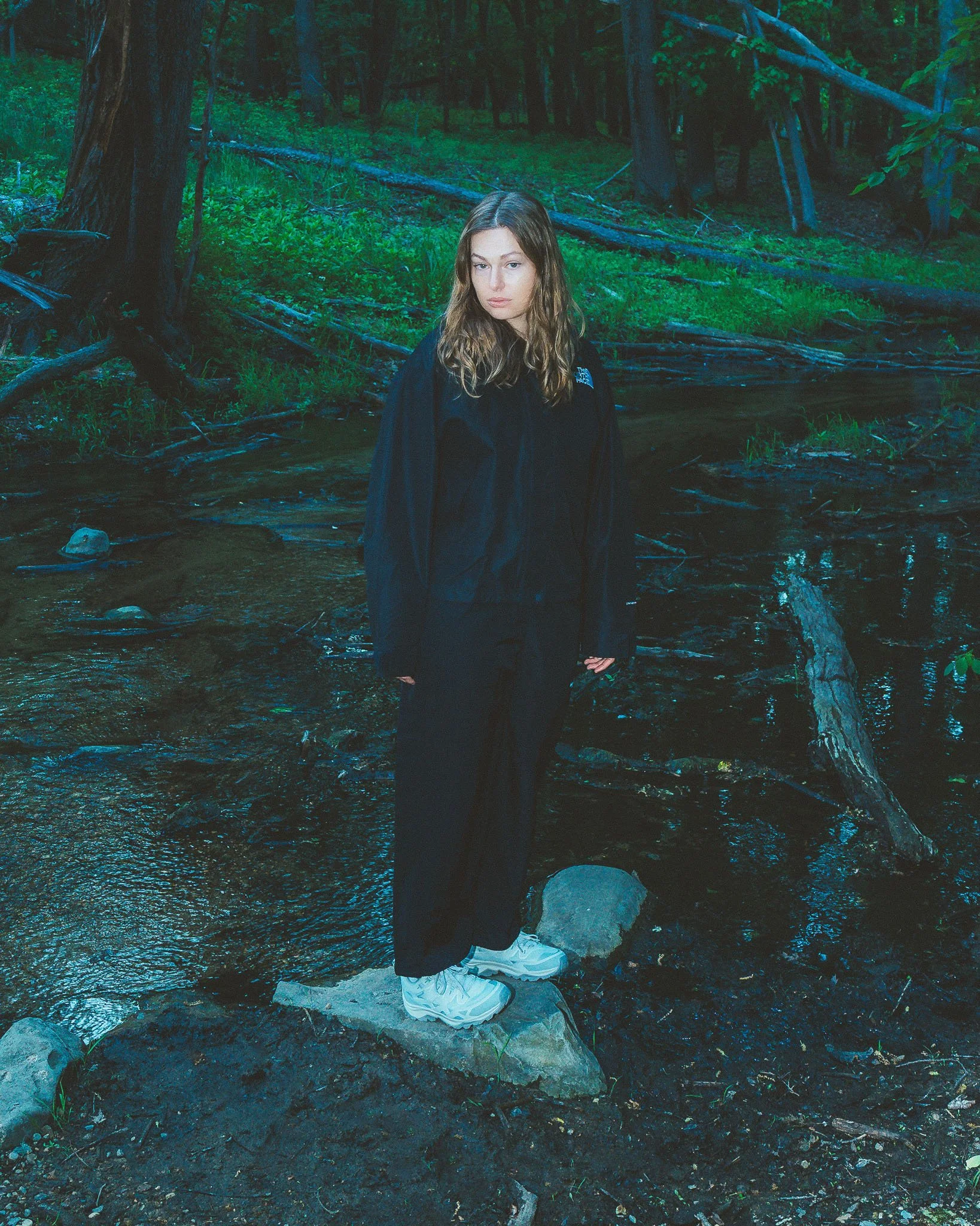 A young woman with wavy, shoulder-length hair stands outdoors in a forested area. She is wearing a black jacket, black pants, and white sneakers. She has a serious expression and is standing on a rocky ground near a small stream.
