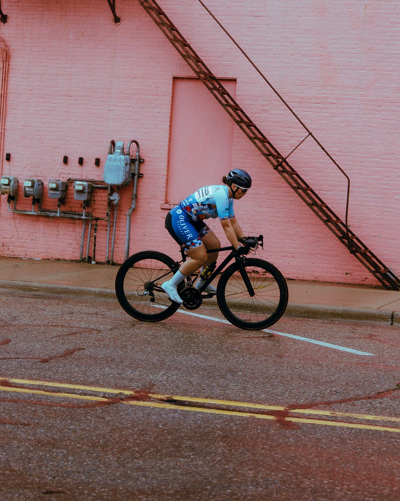 A cyclist in a blue and white uniform riding a black bicycle on a wet street in front of a pink brick wall with a metal fire escape and electrical meters.