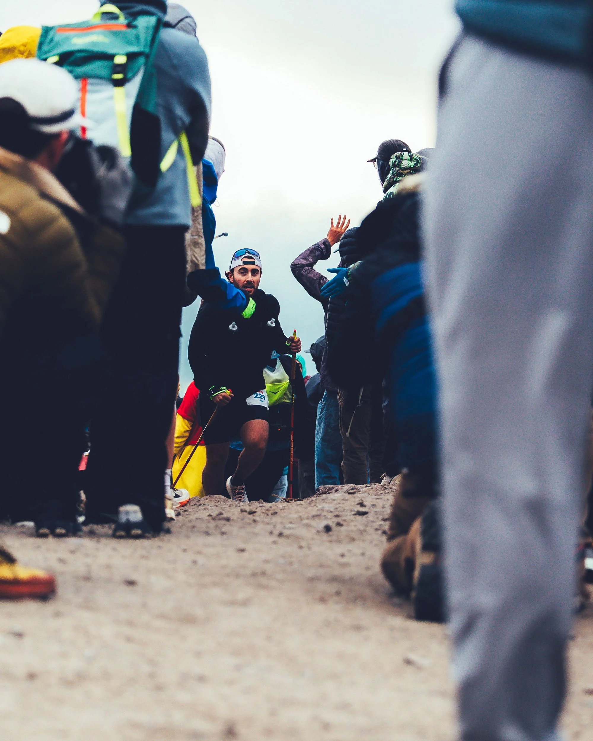 A group of people gathered outdoors, with a focus on a man in the center wearing a blue jacket and hat, holding a trekking pole, and children dressed in athletic clothing, possibly at a hiking or outdoor event.