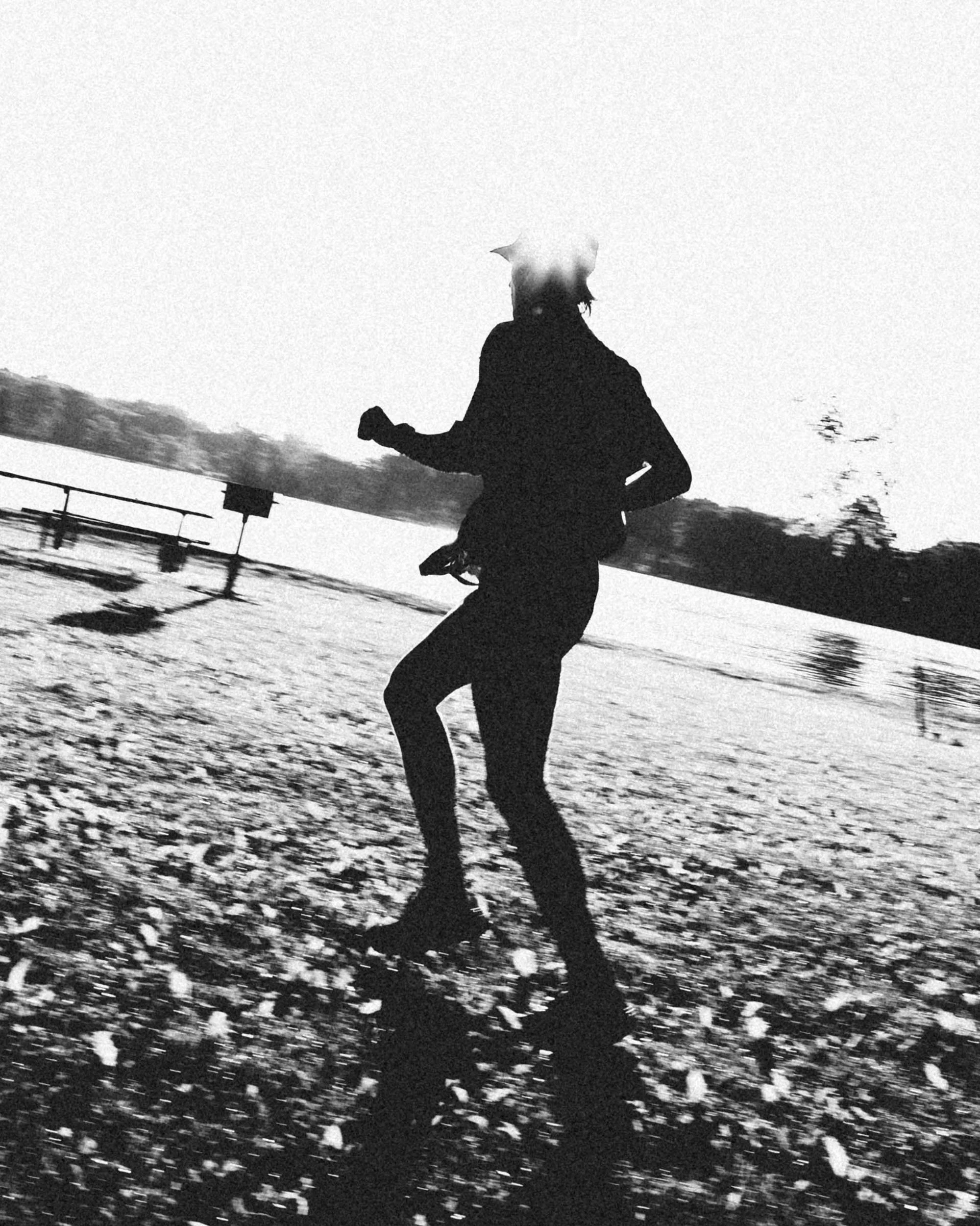 A black and white photo of a person running outside on a snowy ground near a railing, with trees in the background and fog or mist around their head.