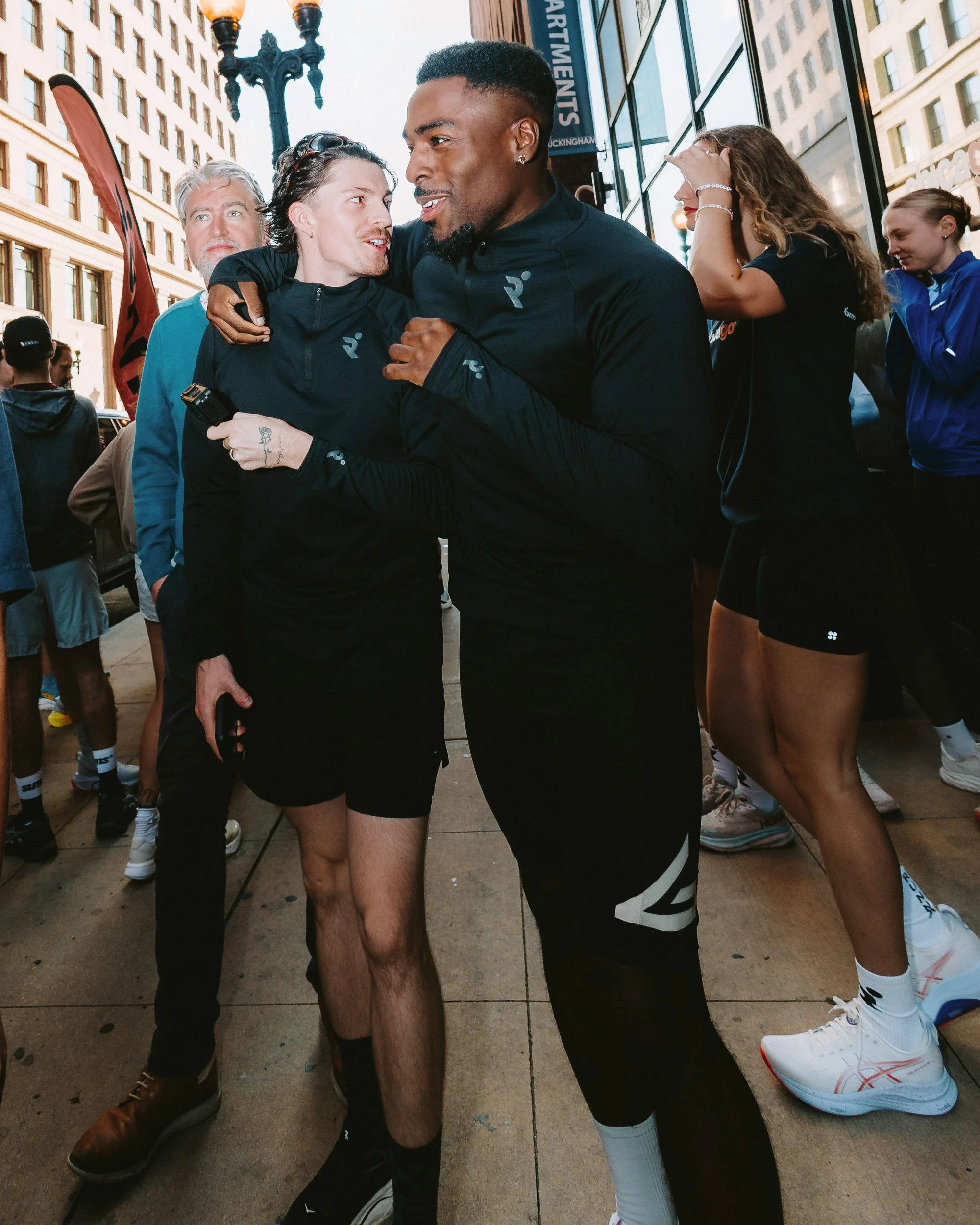 Two men in black athletic jackets embrace and smile at each other on a city sidewalk with tall buildings and a lamppost in the background, surrounded by several women and other people.
