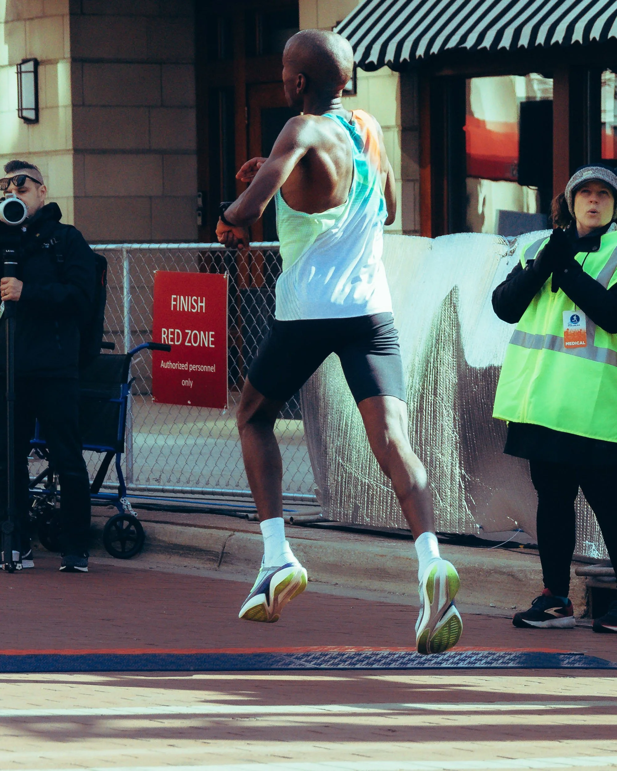 A male runner in mid-air during a race, wearing a white and colorful tank top, black shorts, and white sneakers, with two women and a man standing behind a fence, one of whom is holding a camera and the other wearing a high-visibility vest and medica