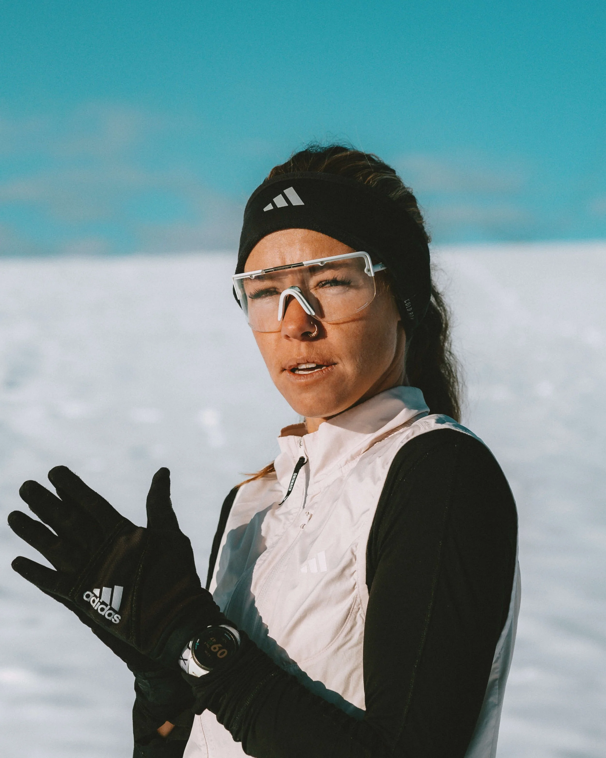 A woman in winter sports gear standing outdoors in a snowy landscape with a clear sky, wearing sunglasses, a black headband, and black gloves, looking into the distance.