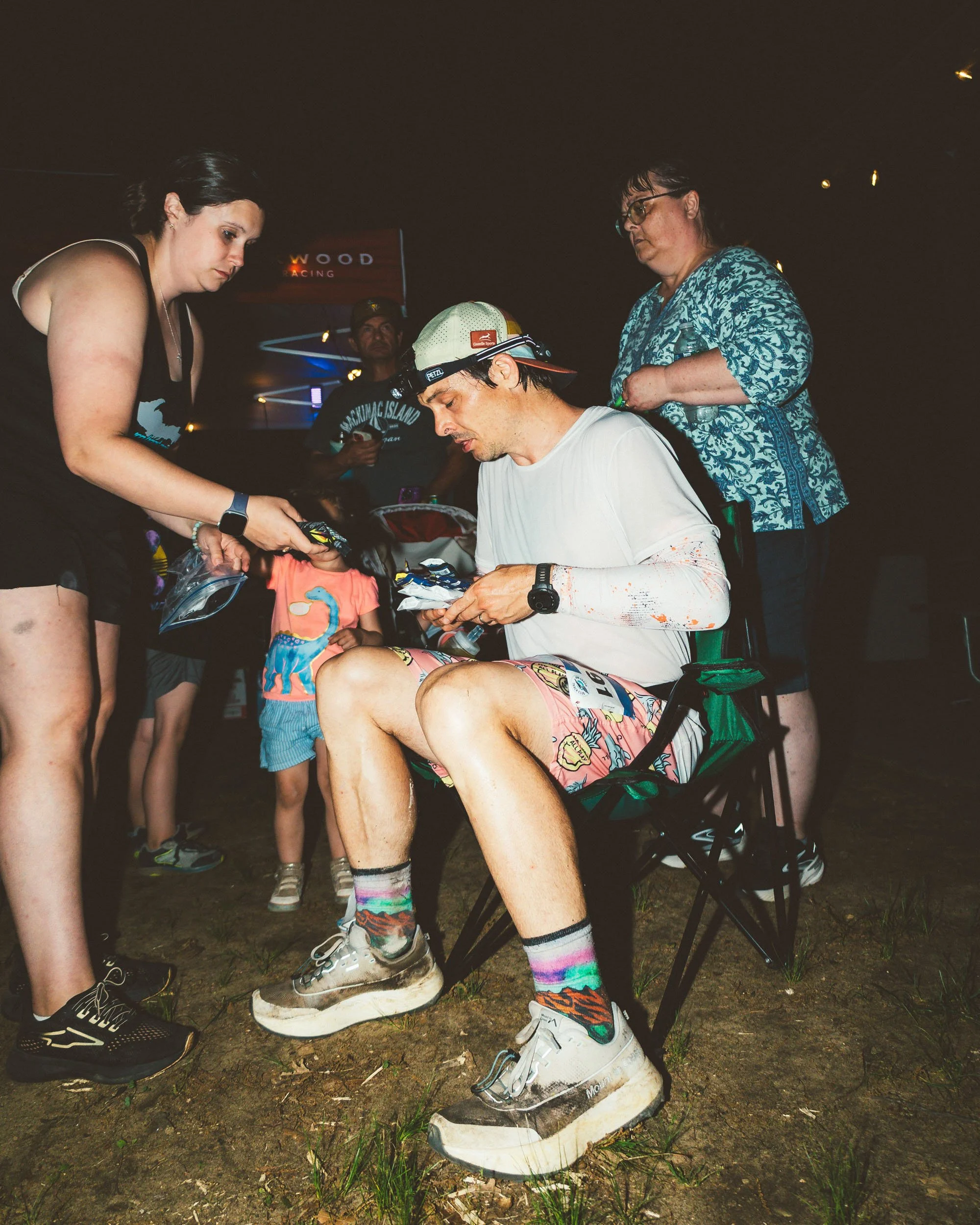 A group of people gathered outdoors at night, with a man sitting in a chair wearing a cap and shorts, surrounded by others standing and tending to him, possibly preparing for a race or event, with a dark background illuminated by lights.