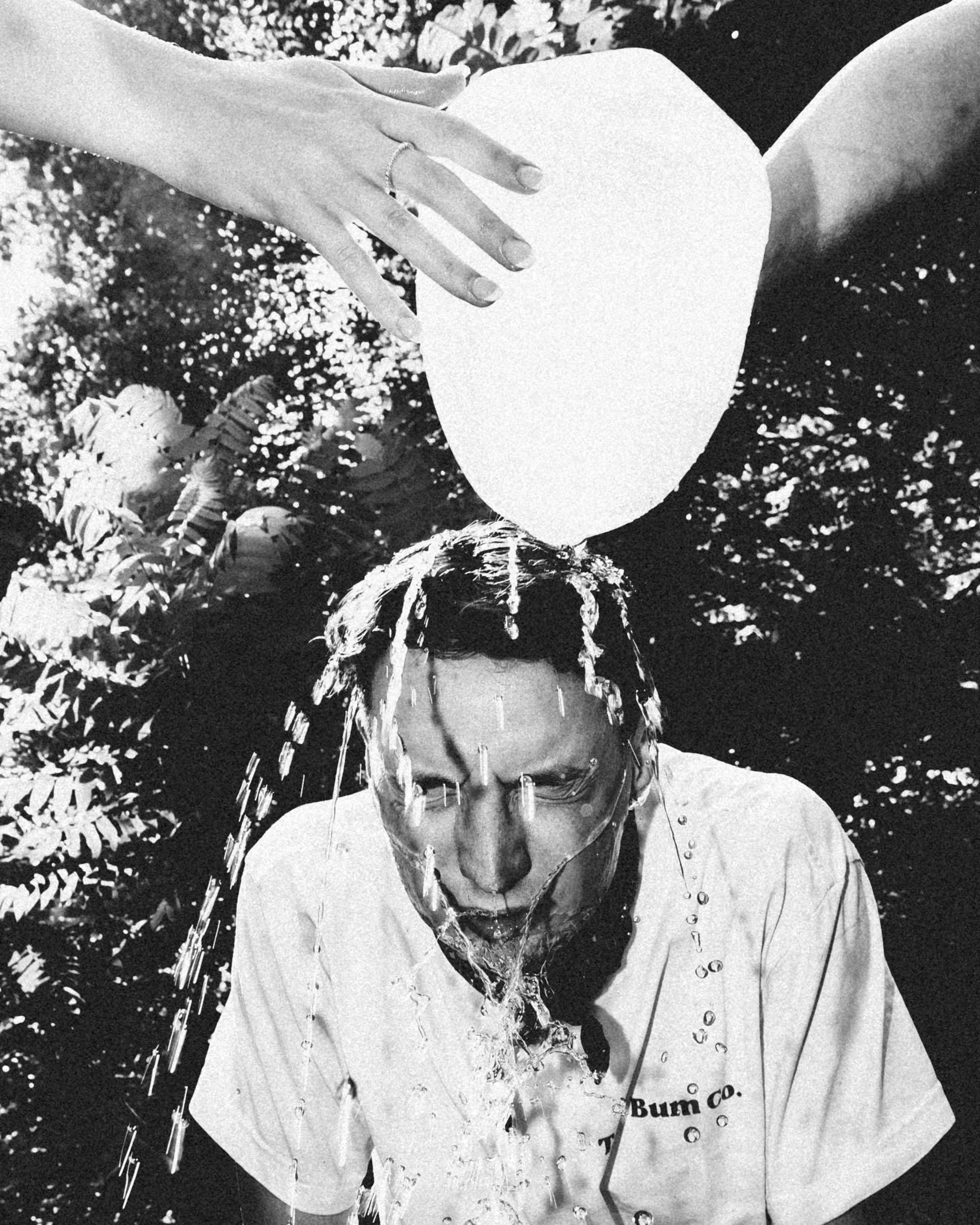 Black and white photo of a woman with her head bowed, pouring a liquid over her hair, with a hand on her forehead and water dripping down. She is outdoors with foliage in the background and is wearing a shirt that has part of the text 'Bum co.' visib