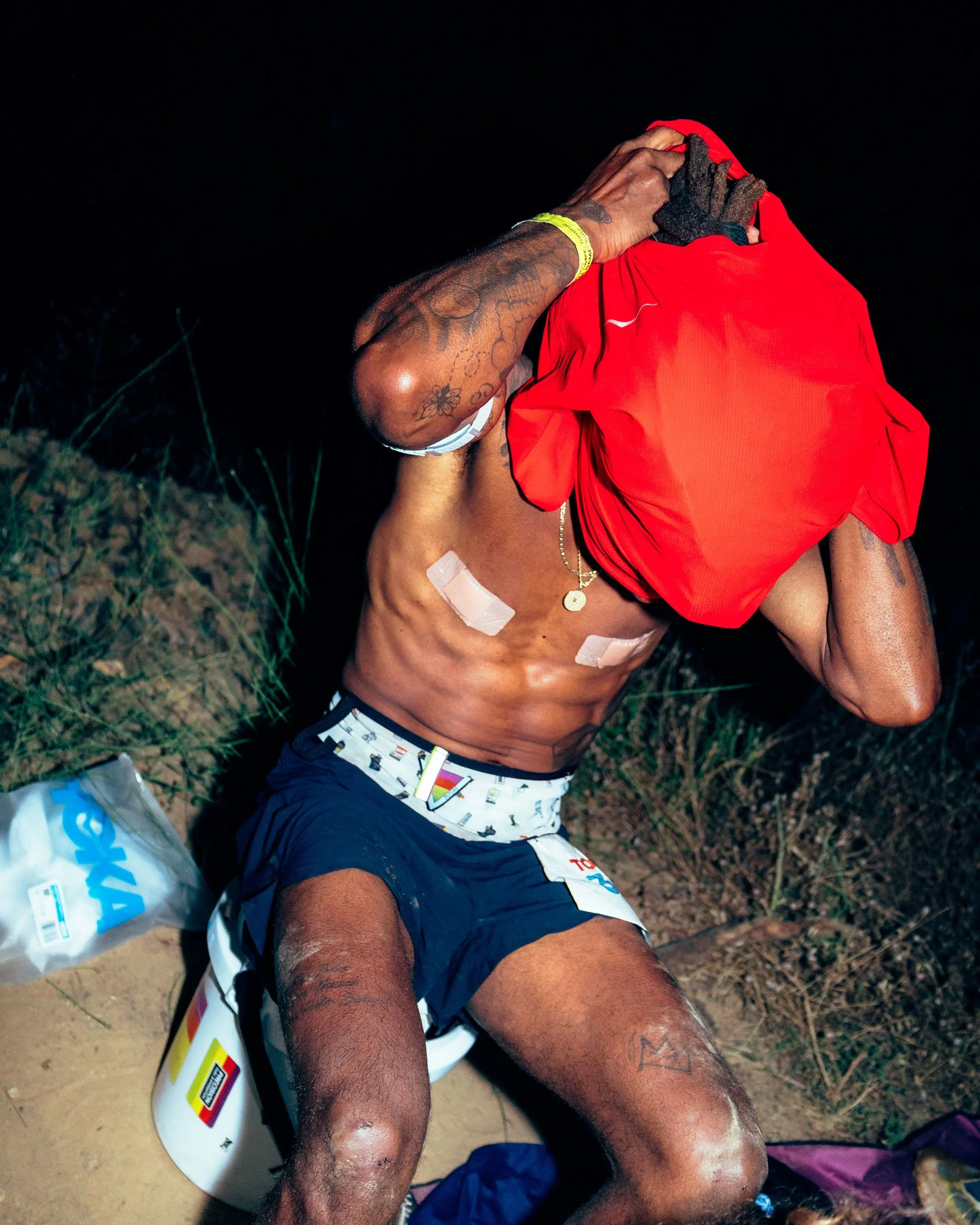 A shirtless man with tattoos on his arms and chest, wearing swimming shorts, sitting outdoors at night, holding a red cloth or shirt over his head, with bandages on his arms and legs.