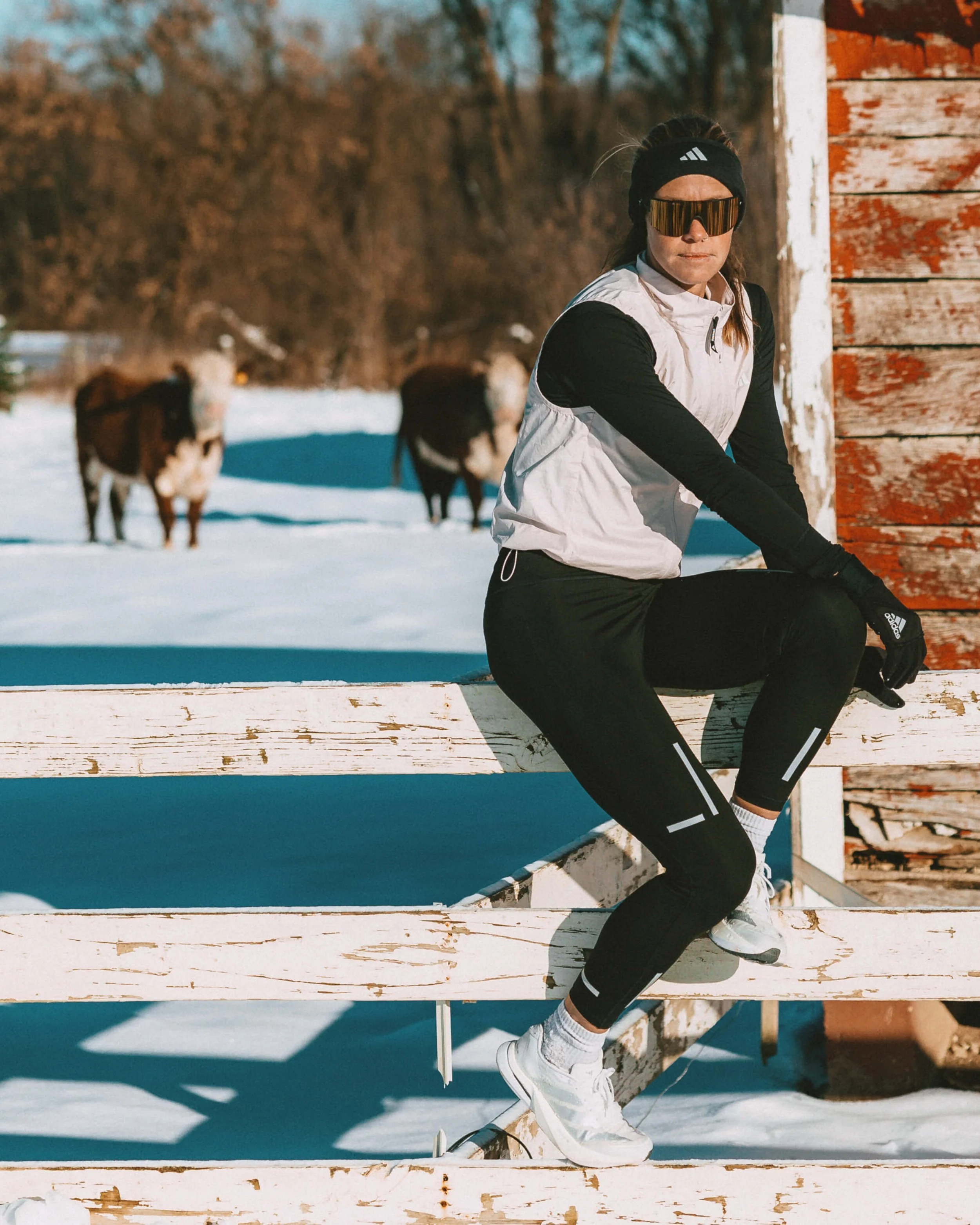 A woman in athletic gear, including sunglasses, a headband, a white and black jacket, and black leggings, sitting on a snowy wooden structure outdoors with trees and animals in the background.