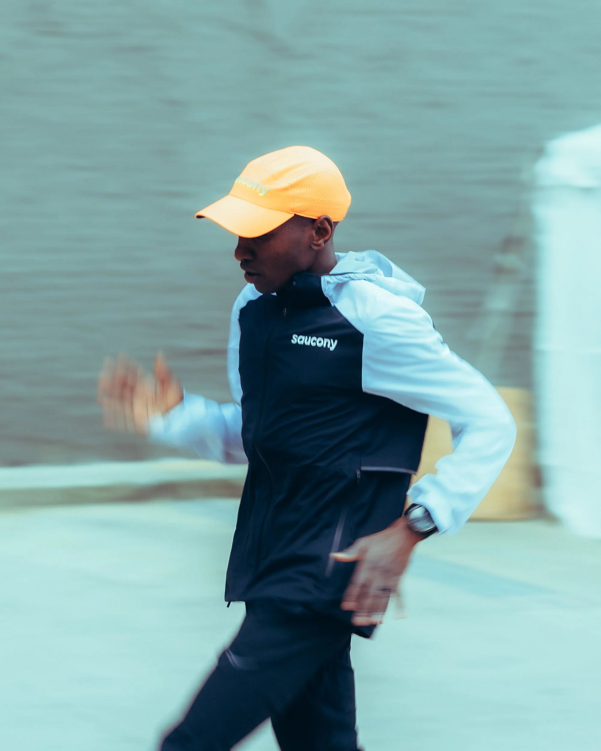Person wearing an orange cap and black Saucony jacket running near a body of water.