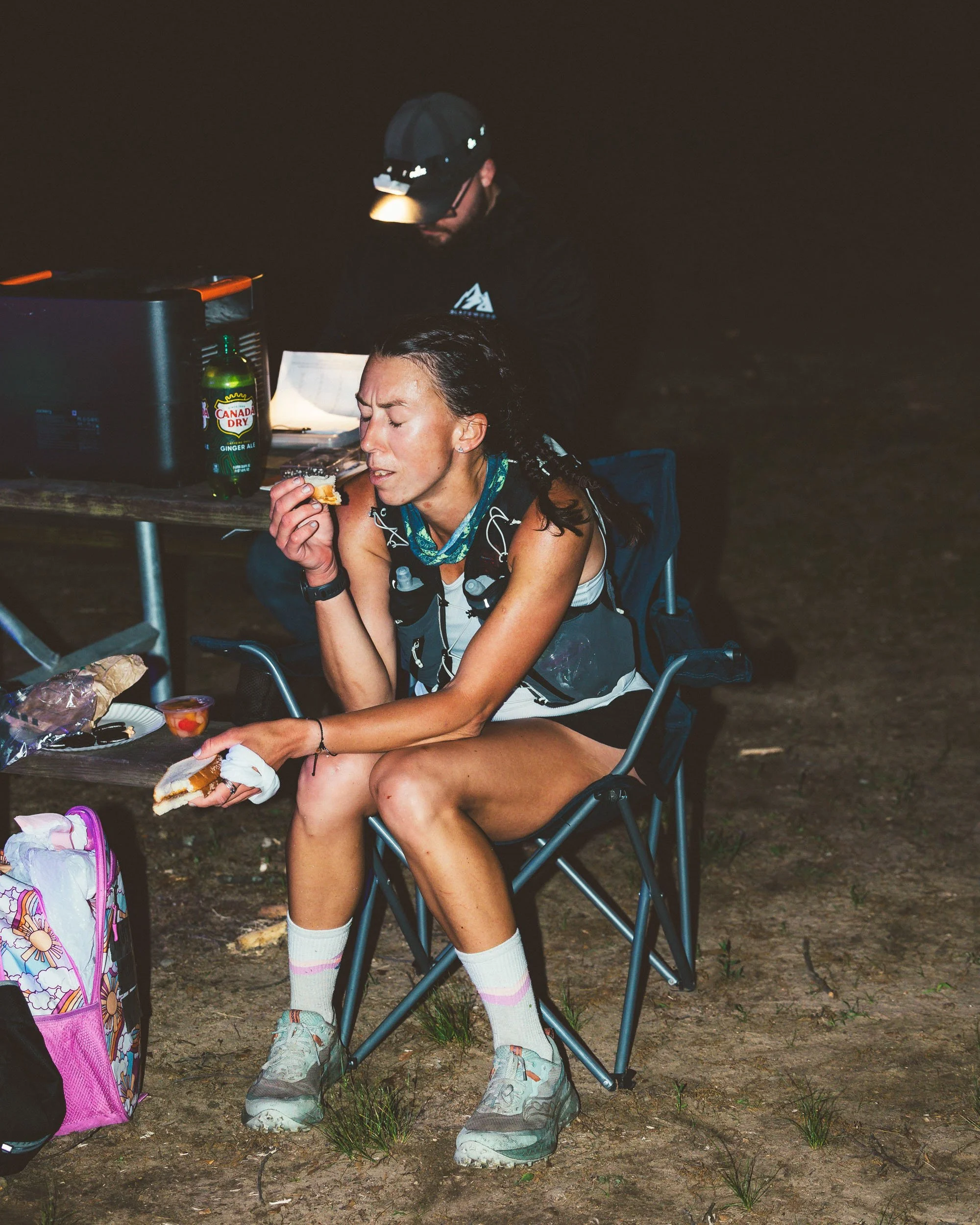 A woman with dark hair in a braid, wearing a tank top, socks, and running shoes, is sitting on a folding chair at night, eating a piece of food. She is outdoors on dirt ground with small grass patches. Behind her, a man in a cap and dark clothing is 