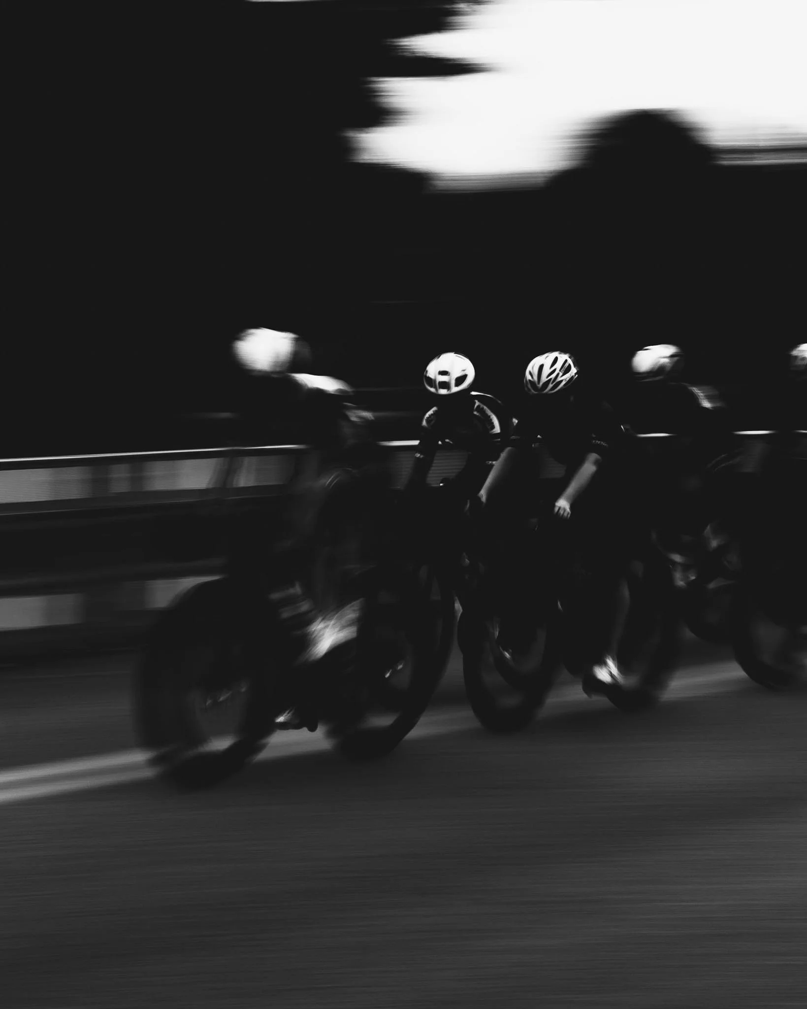 A group of cyclists riding bicycles on a road, with motion blur indicating speed, wearing helmets and cycling gear.