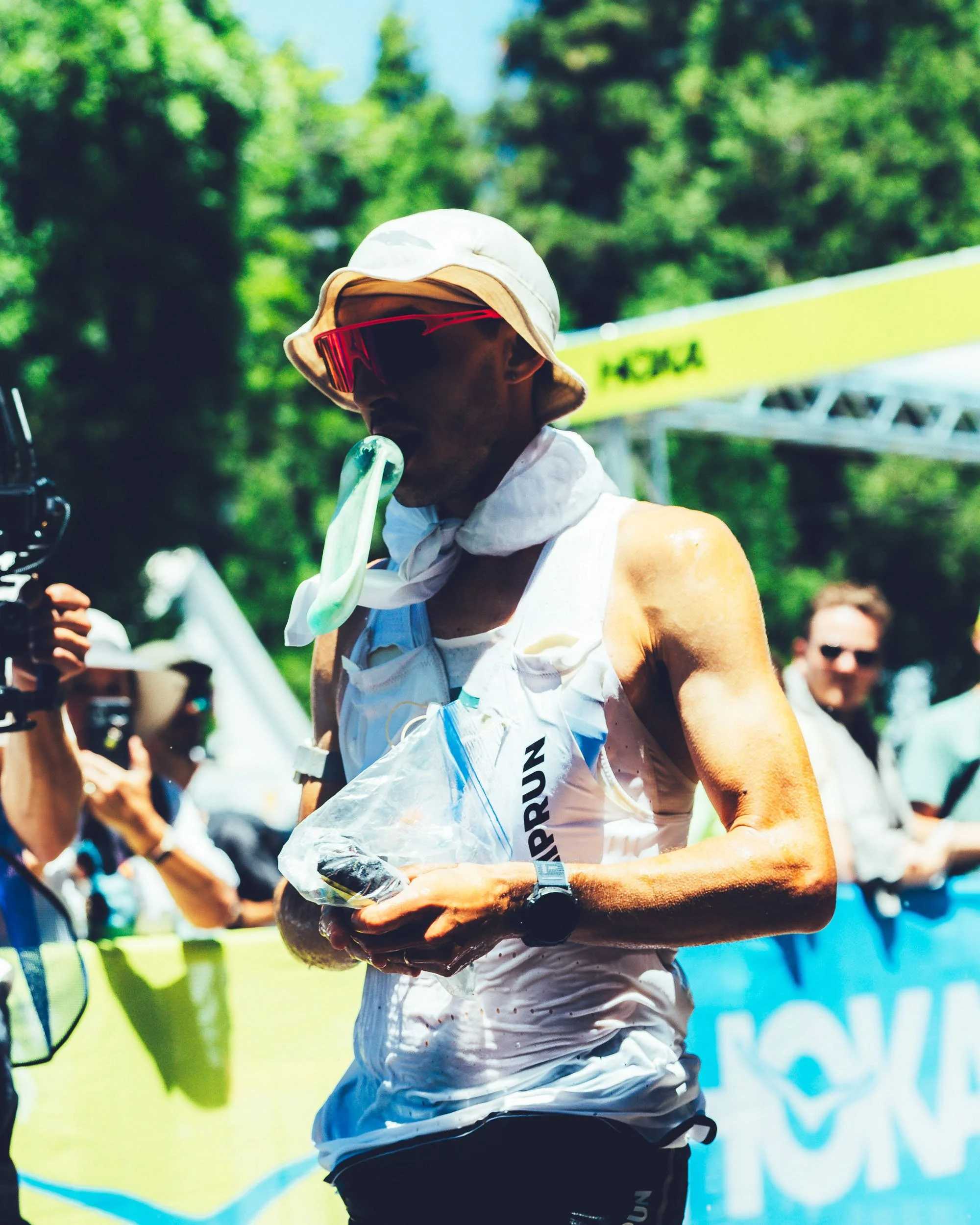 A person participating in a race wearing a white hat, red sunglasses, and a race bib, with a green and blue background and a yellow signage in the background.
