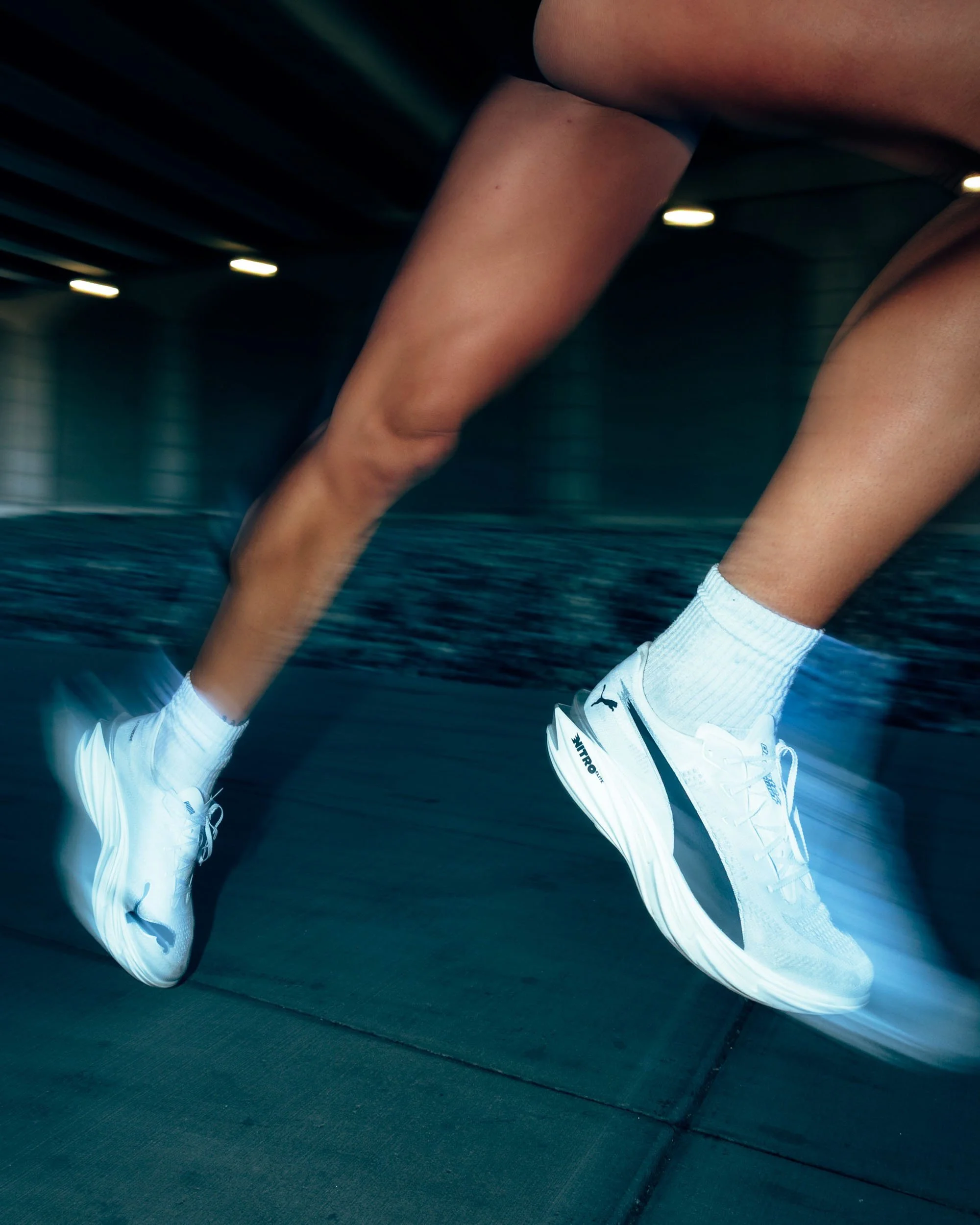 Close-up of a person's legs in white sports shoes and socks, positioned against a dark surface, with a dark background and ceiling lights visible.