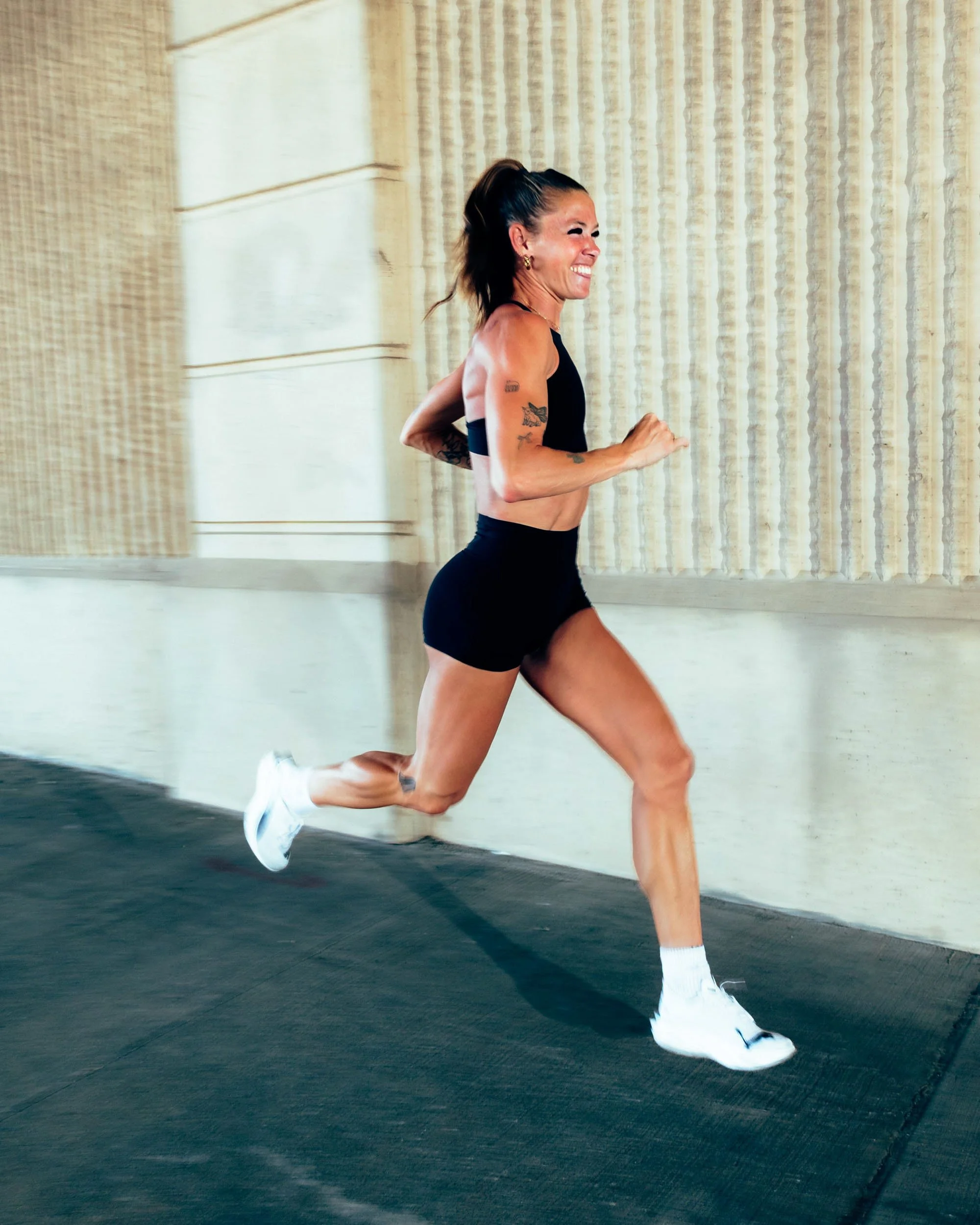 A woman running indoors, dressed in athletic black shorts and a black sports bra, wearing white sneakers, smiling as she exercises on a black floor with a beige wall in the background.