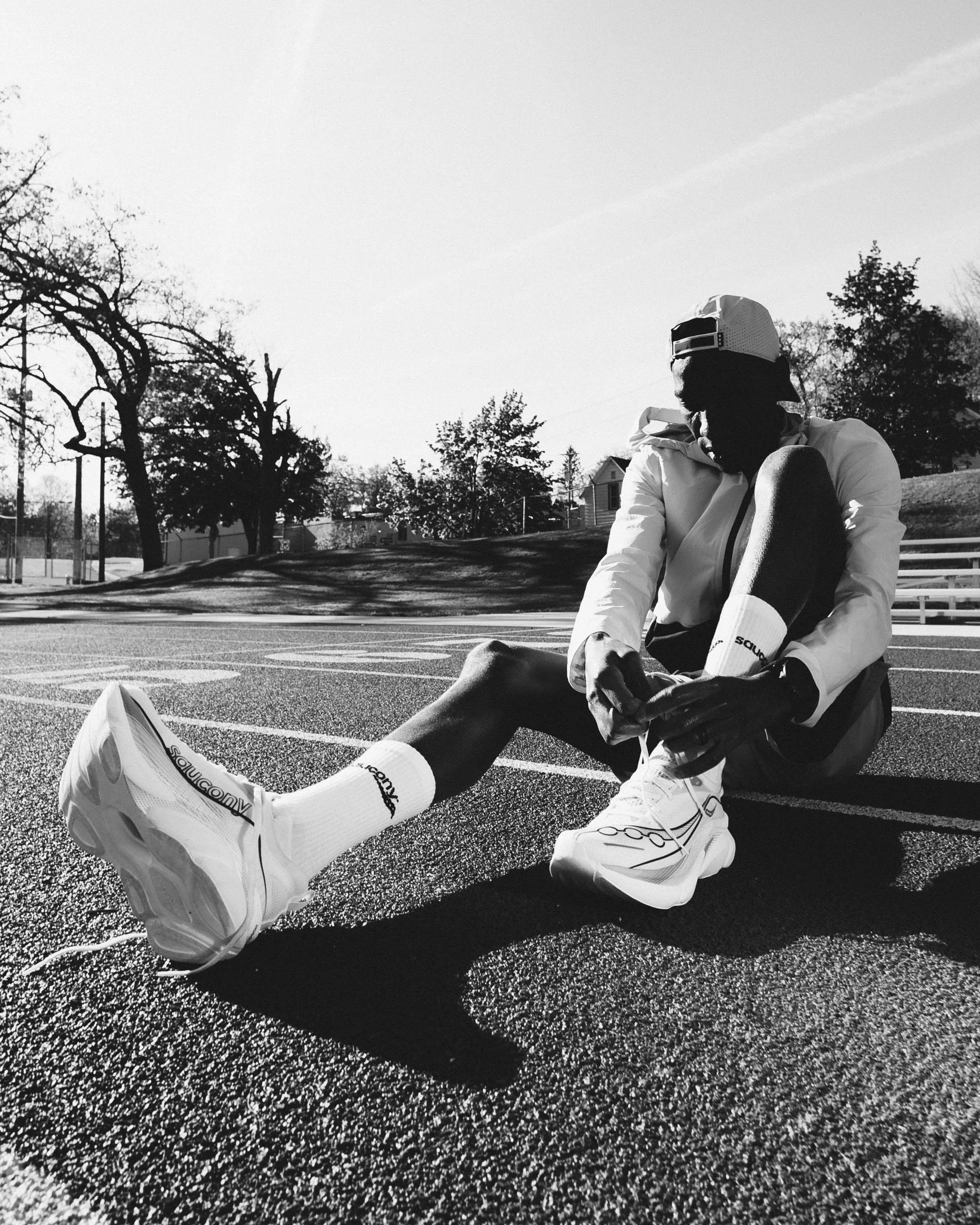 A person sitting on a running track, wearing running shoes, athletic clothing, and a cap, with trees and a fence in the background.