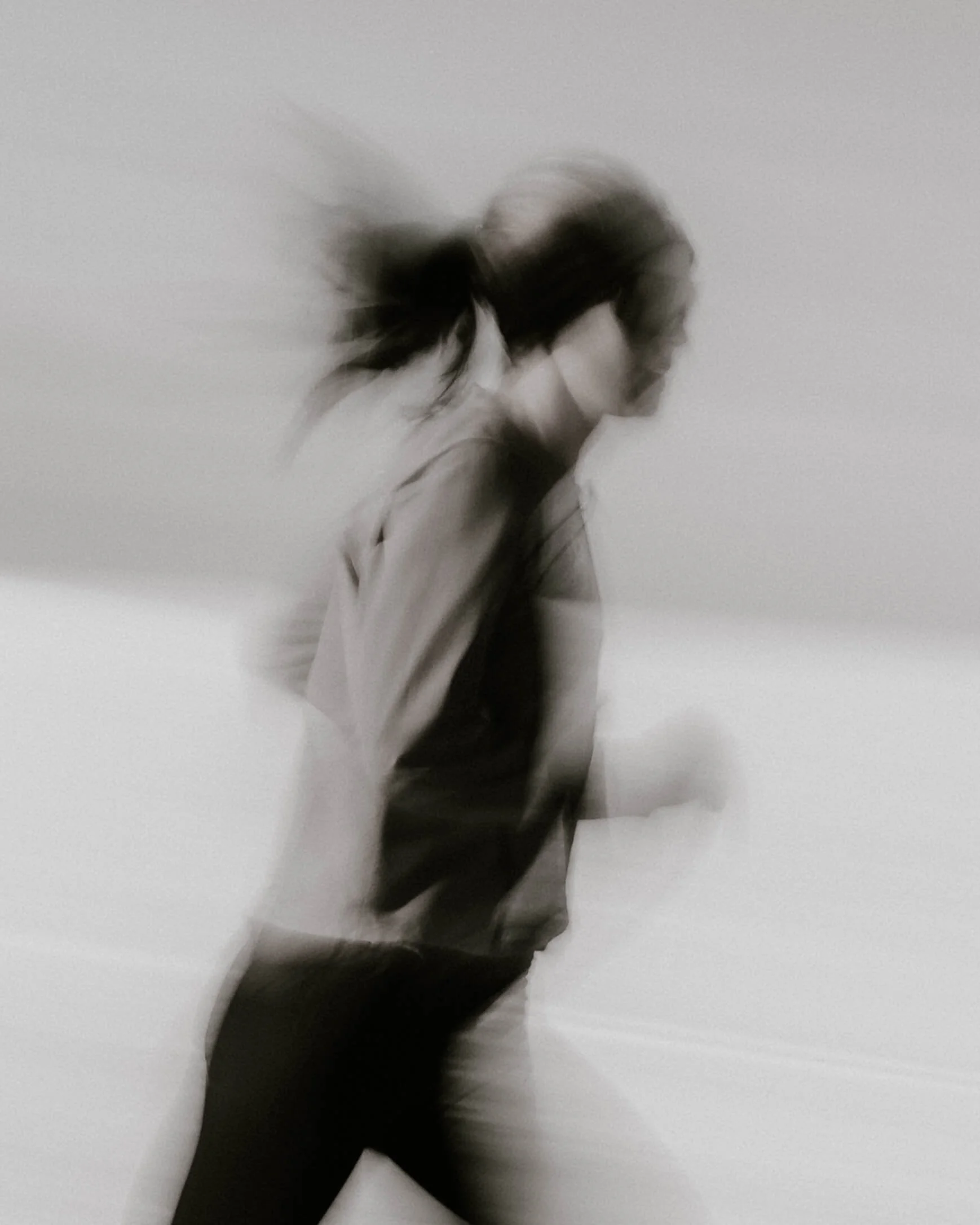 A blurry black and white photo of a person with long hair walking in a white room.